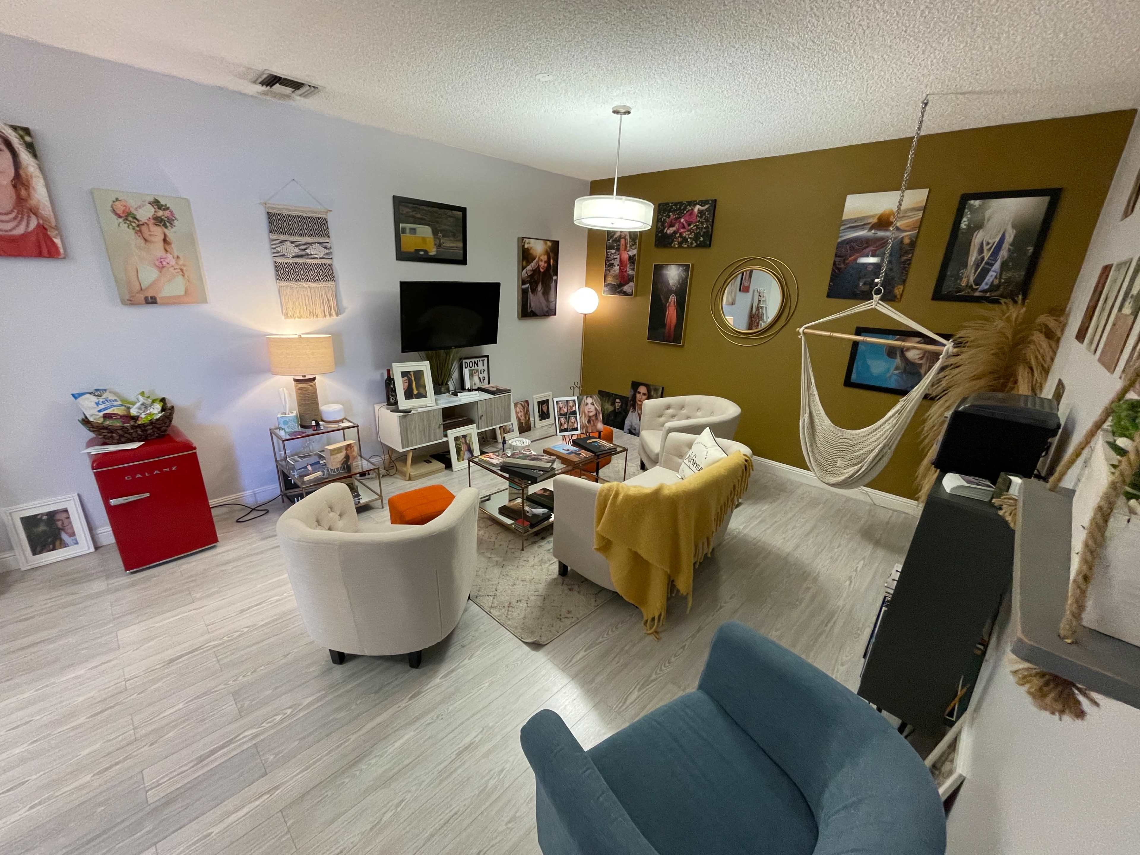 The room features a mix of seating options, a decorative wall with framed photos, and a small red refrigerator in one corner.
