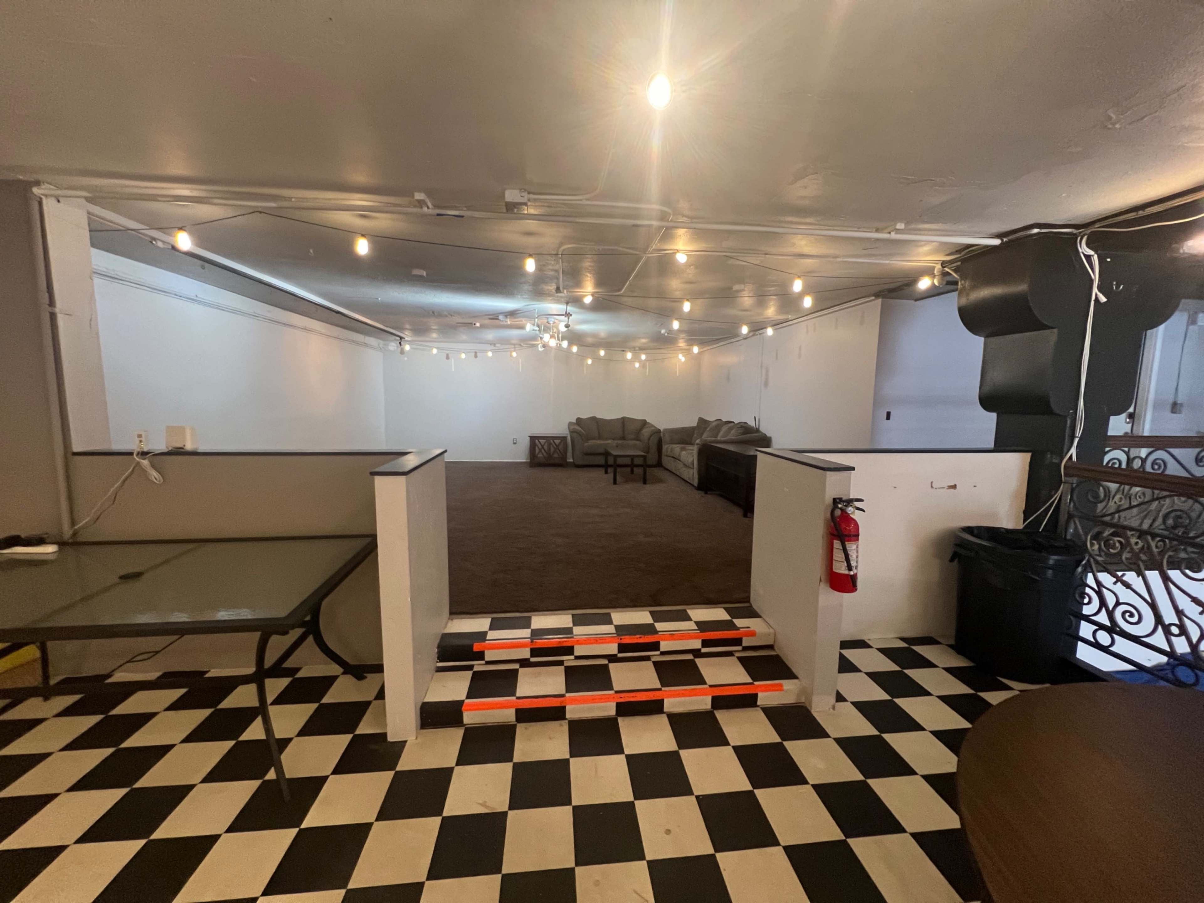 The image shows a spacious, empty room with a checkered black and white floor, string lights overhead, and seating arranged against the walls.