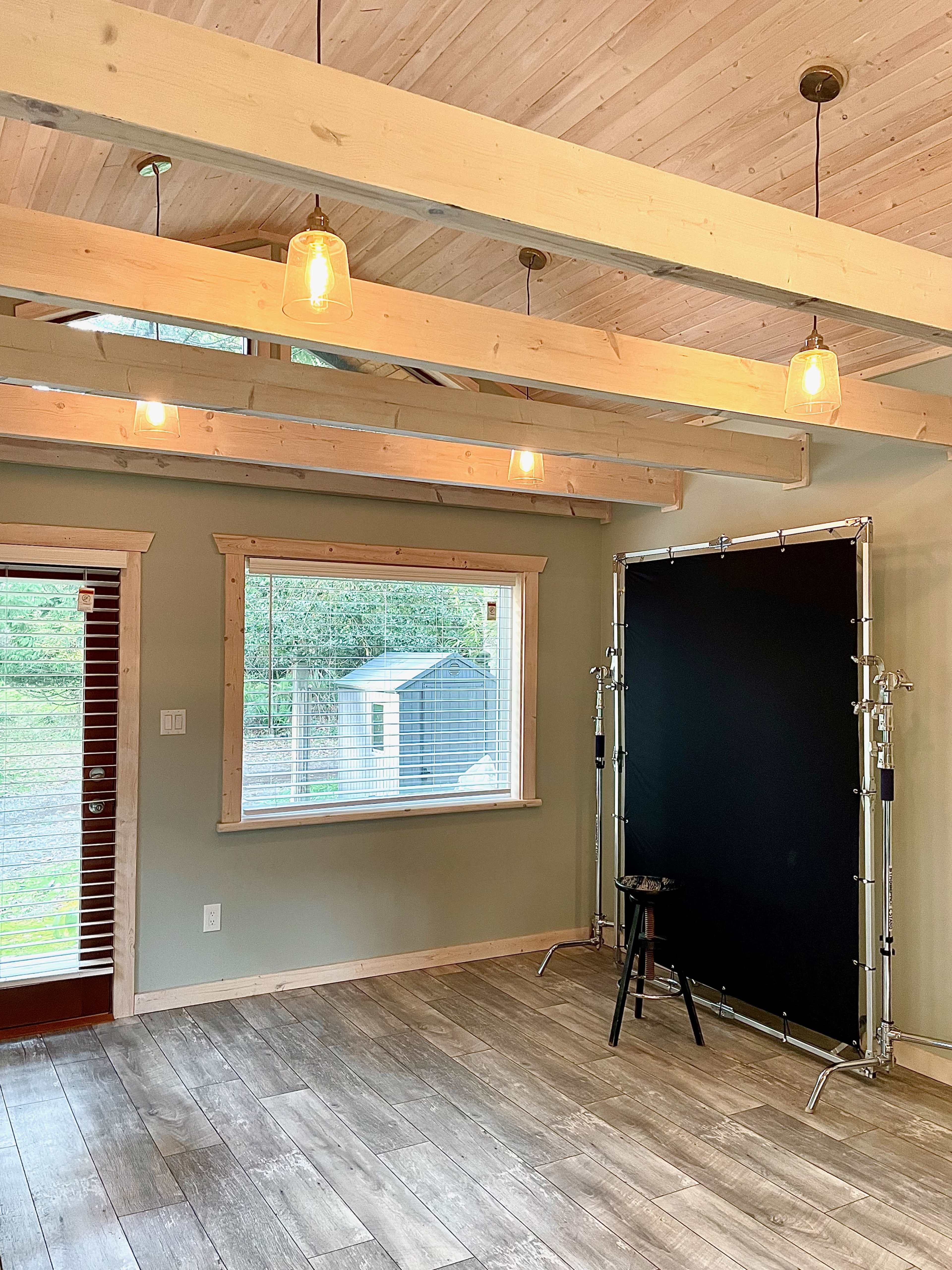 A well-lit room with wooden beams features a black backdrop and a stool, along with a window overlooking a garden area.