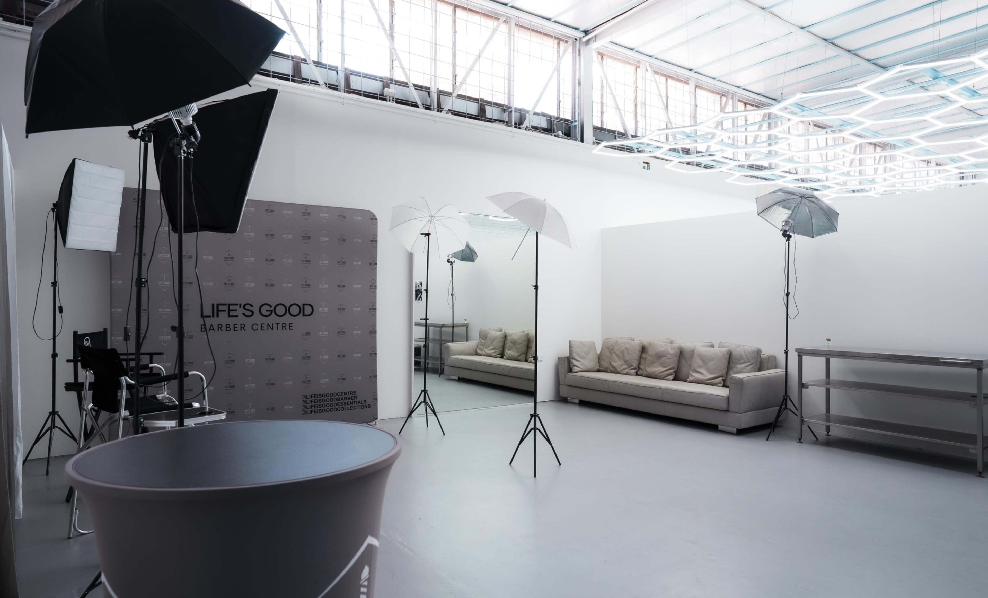 The image shows a well-lit barber studio featuring a gray sofa, a large mirror, and several photography lights set up in the space.