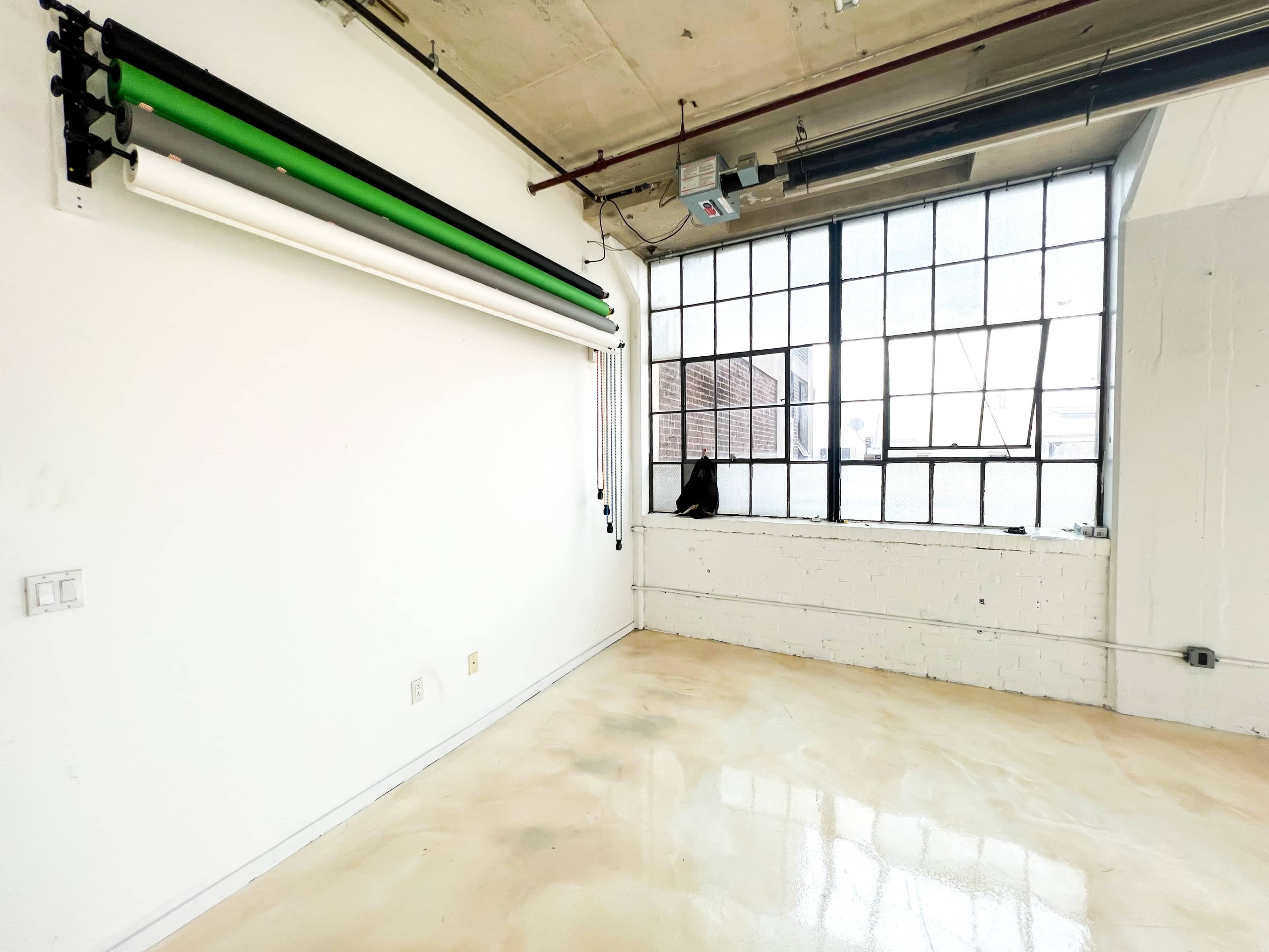 A bright, empty room with a large window and a green backdrop setup on the wall.
