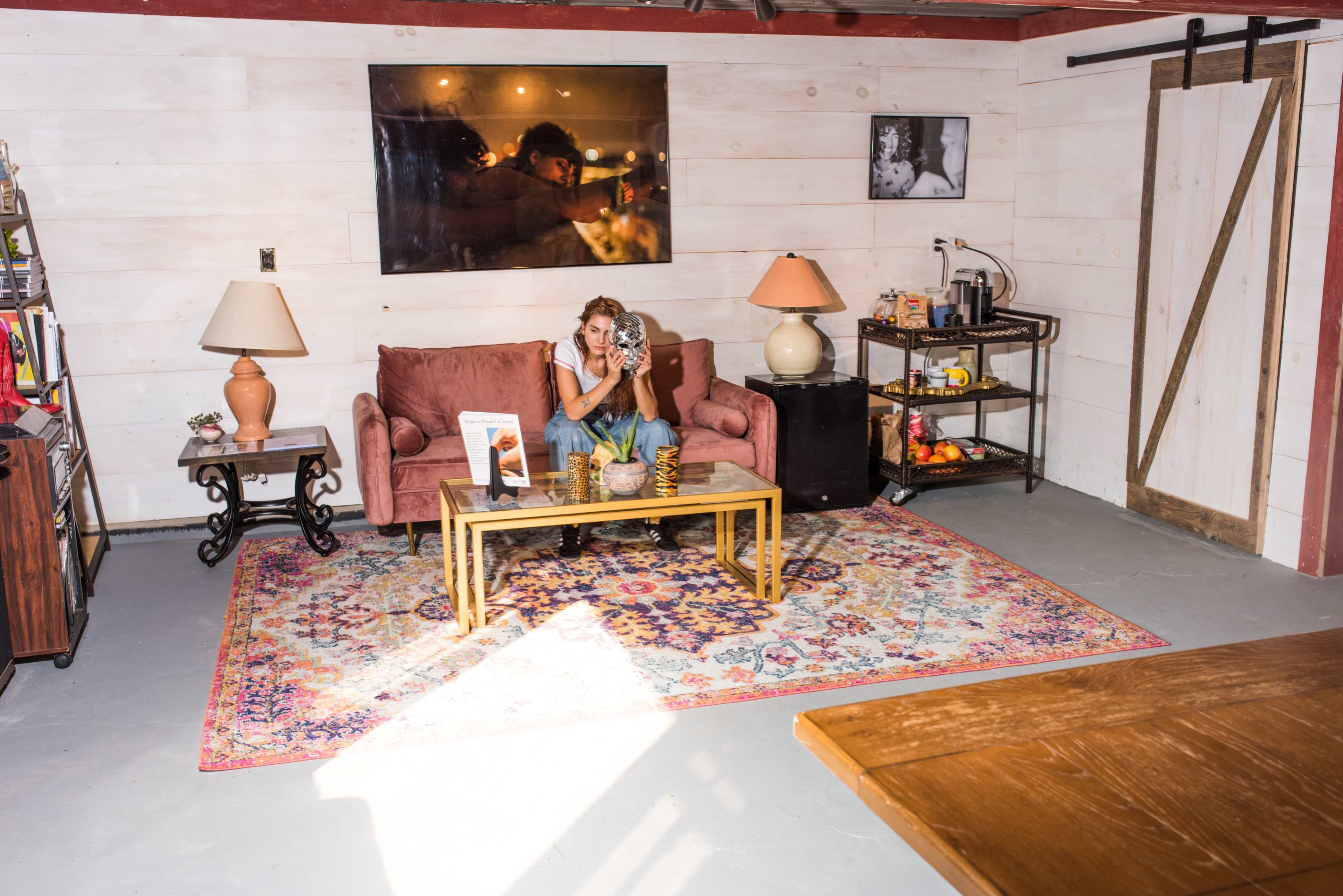 A woman sits on a pink couch in a rustic room adorned with a large rug, a table in front of her, and various decorative items around.