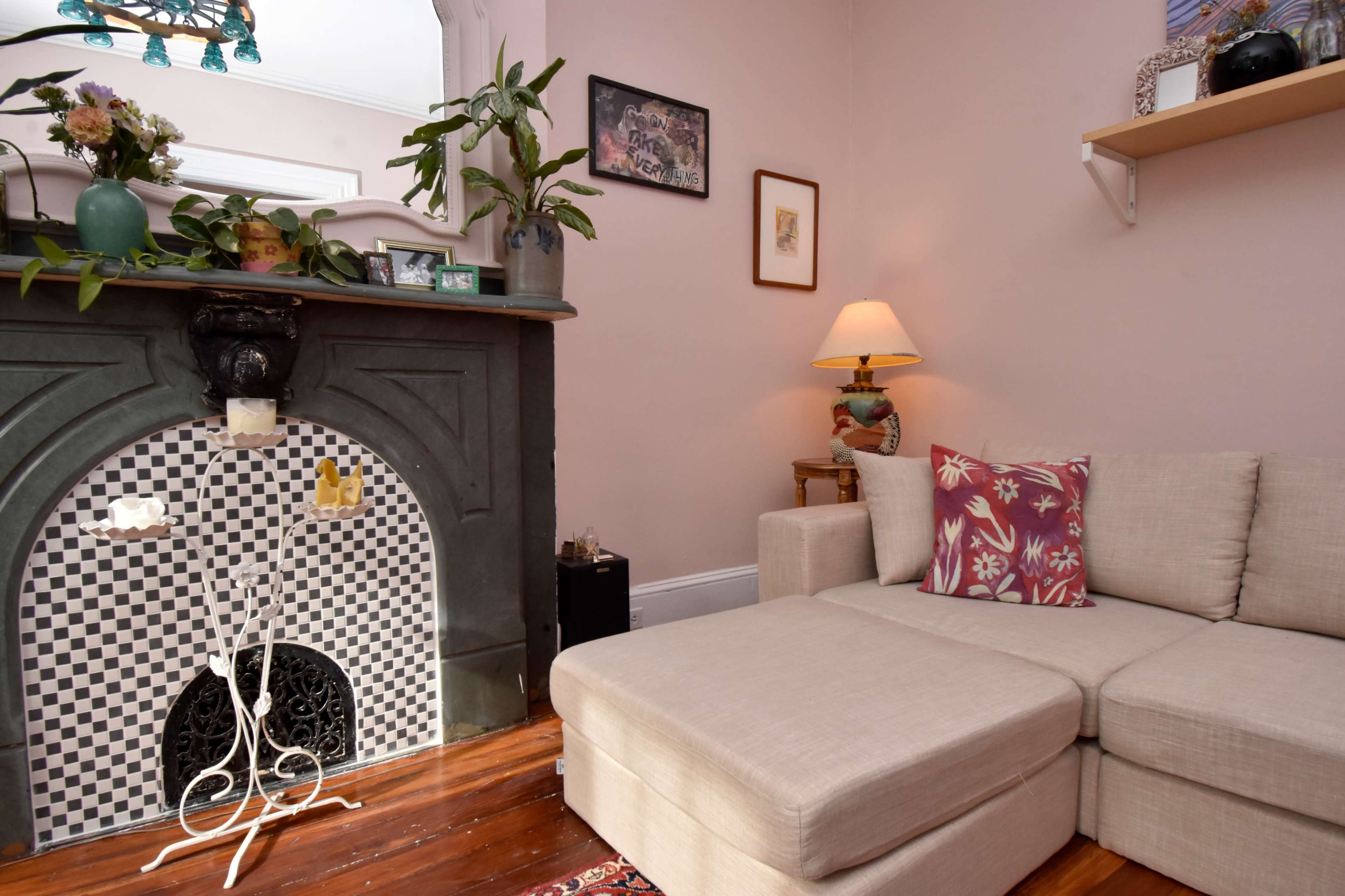 The image shows a cozy living room with a light-colored sectional sofa, an ornate fireplace with a black and white tiled design, and a small lamp on a side table.