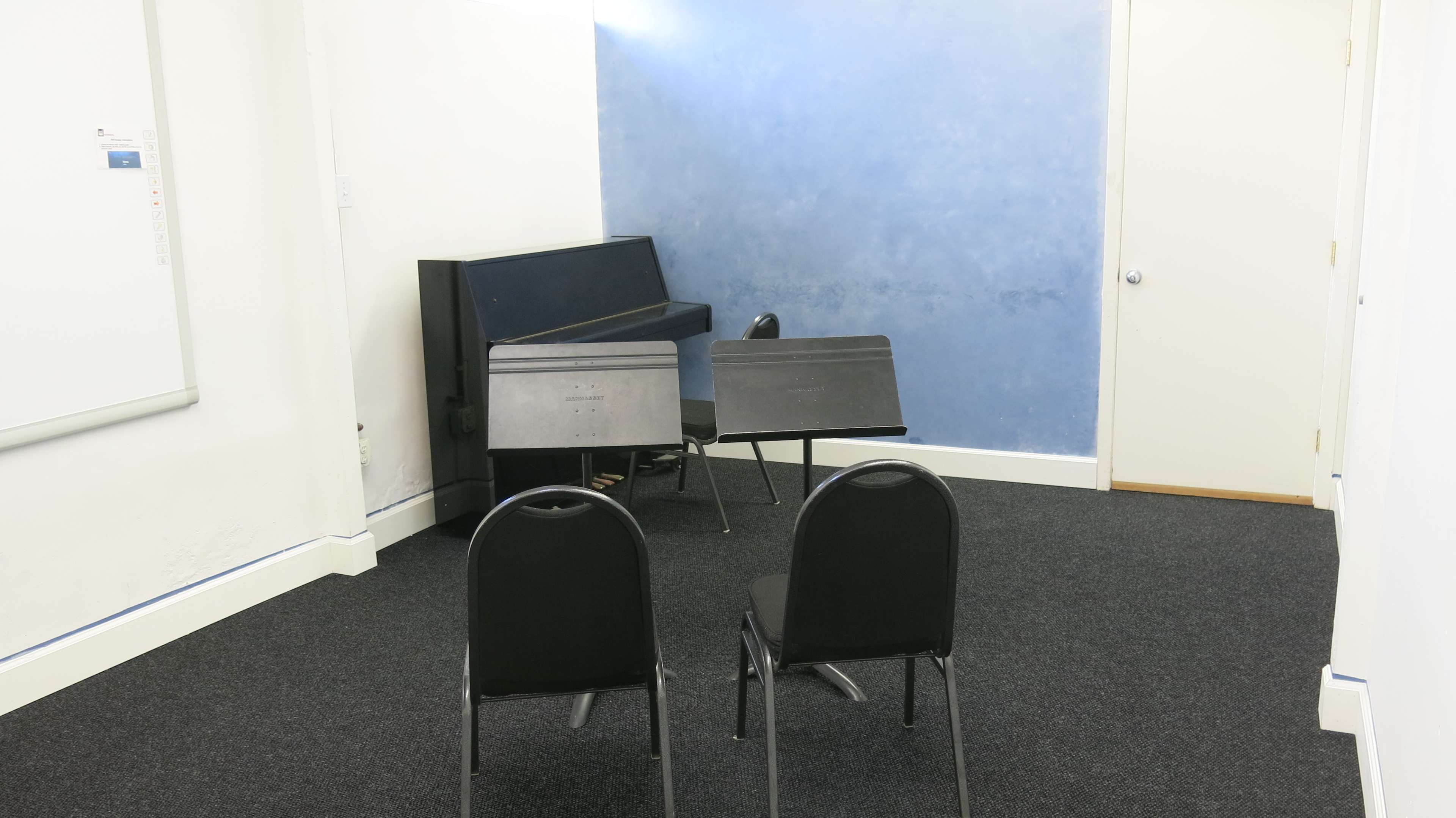 The image shows a small room with two black chairs facing a blue wall, a stacked piano, and music stands in the corner.