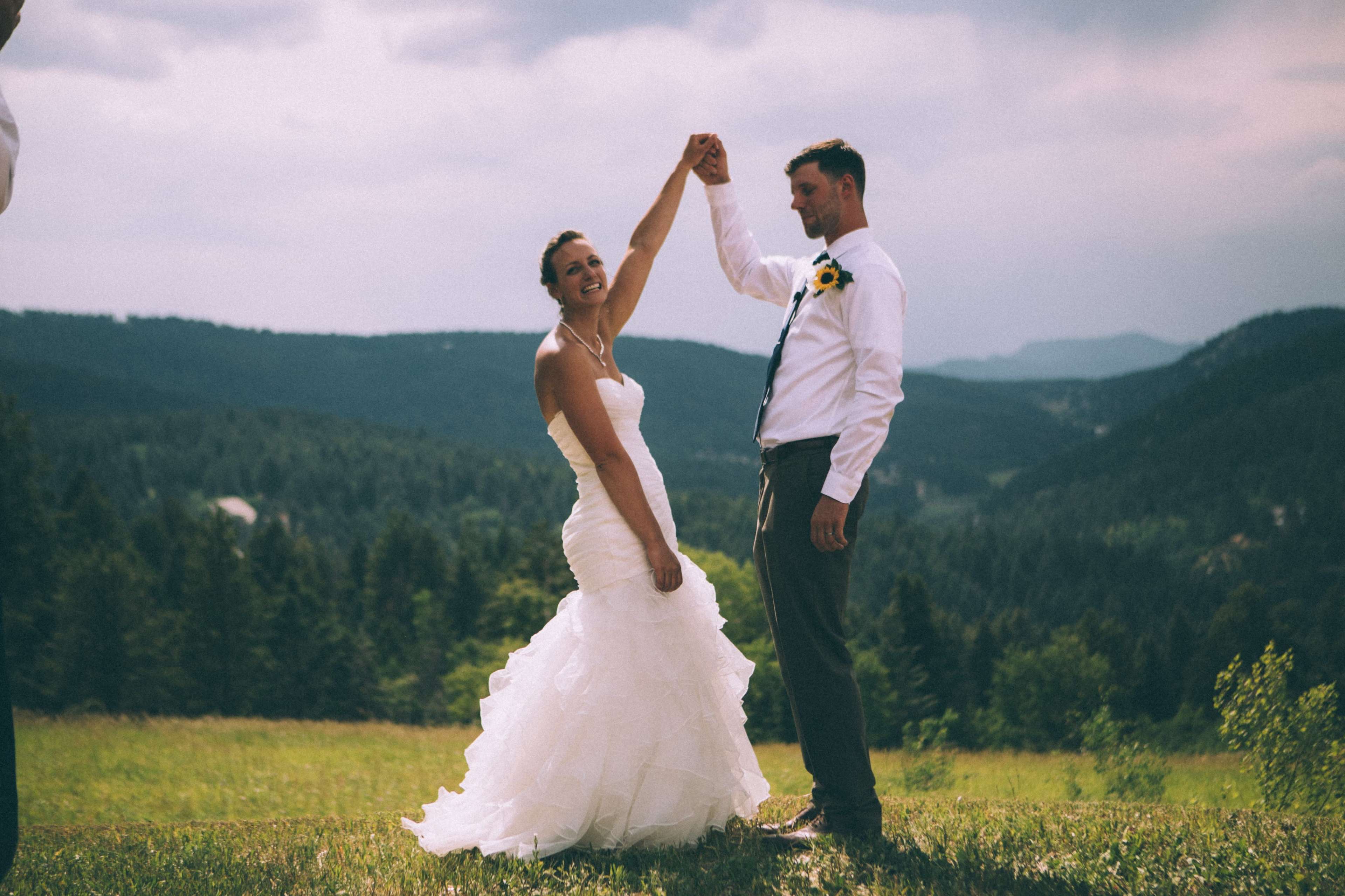 A bride and groom dance outdoors, holding hands while surrounded by a forested landscape and distant mountains.