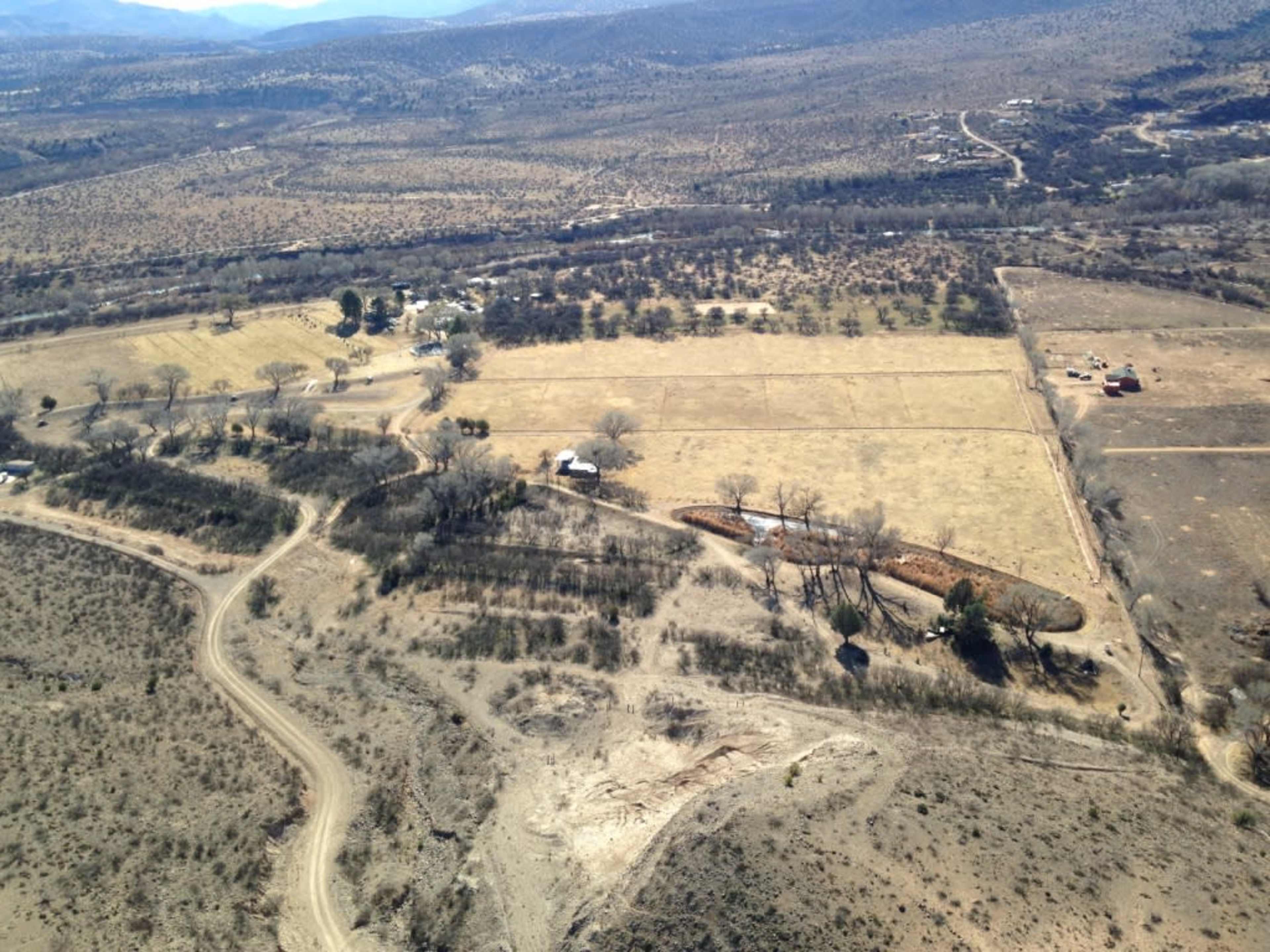 The image shows a vast landscape with a mix of open fields, patches of vegetation, and winding dirt roads under a clear sky.