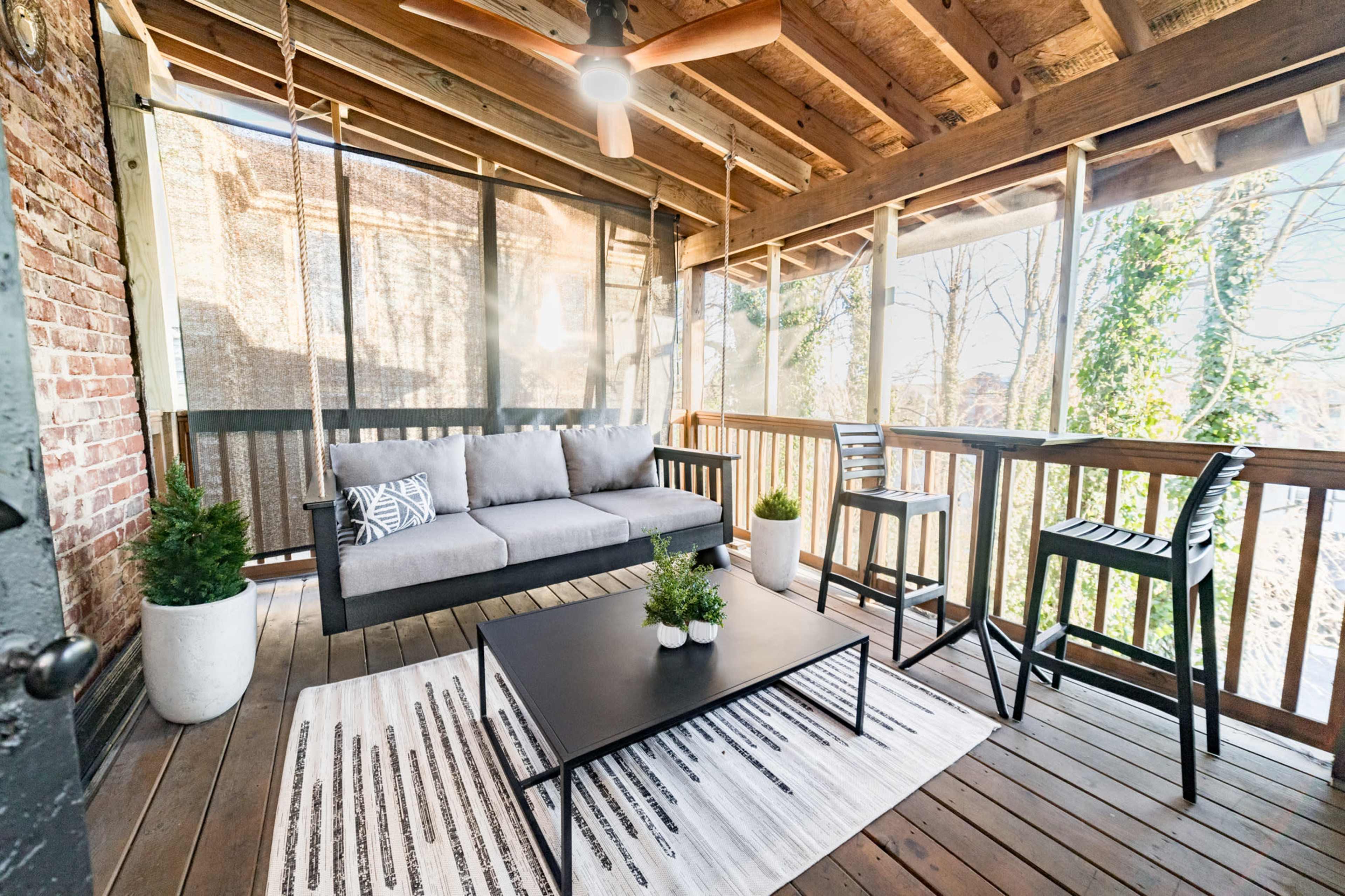 A covered outdoor patio features a gray sofa, a black coffee table, and two high chairs, surrounded by greenery and wooden beams.
