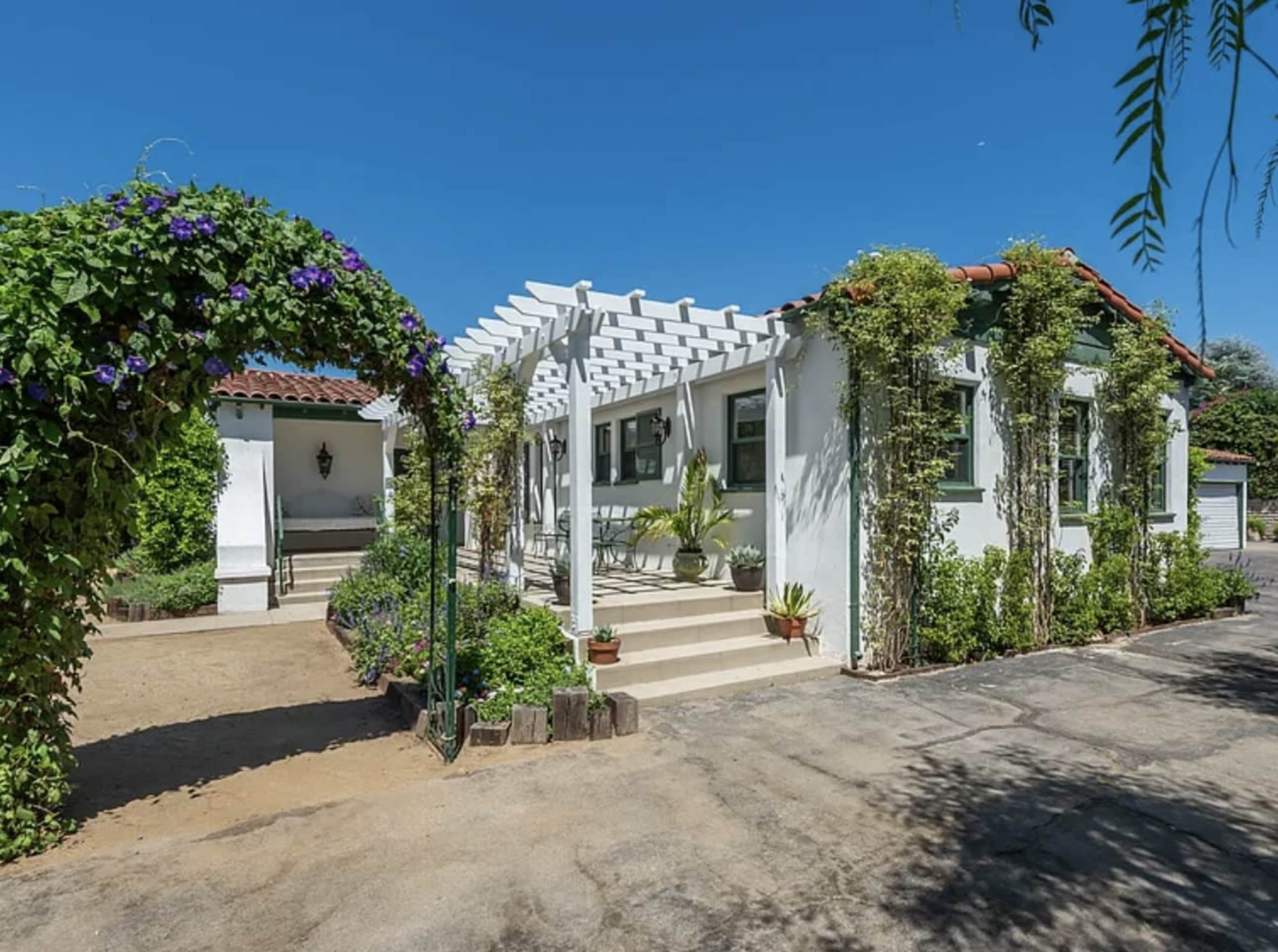 The image shows a white house with a pergola covered in greenery, located beside a gravel pathway and surrounded by a landscaped yard.