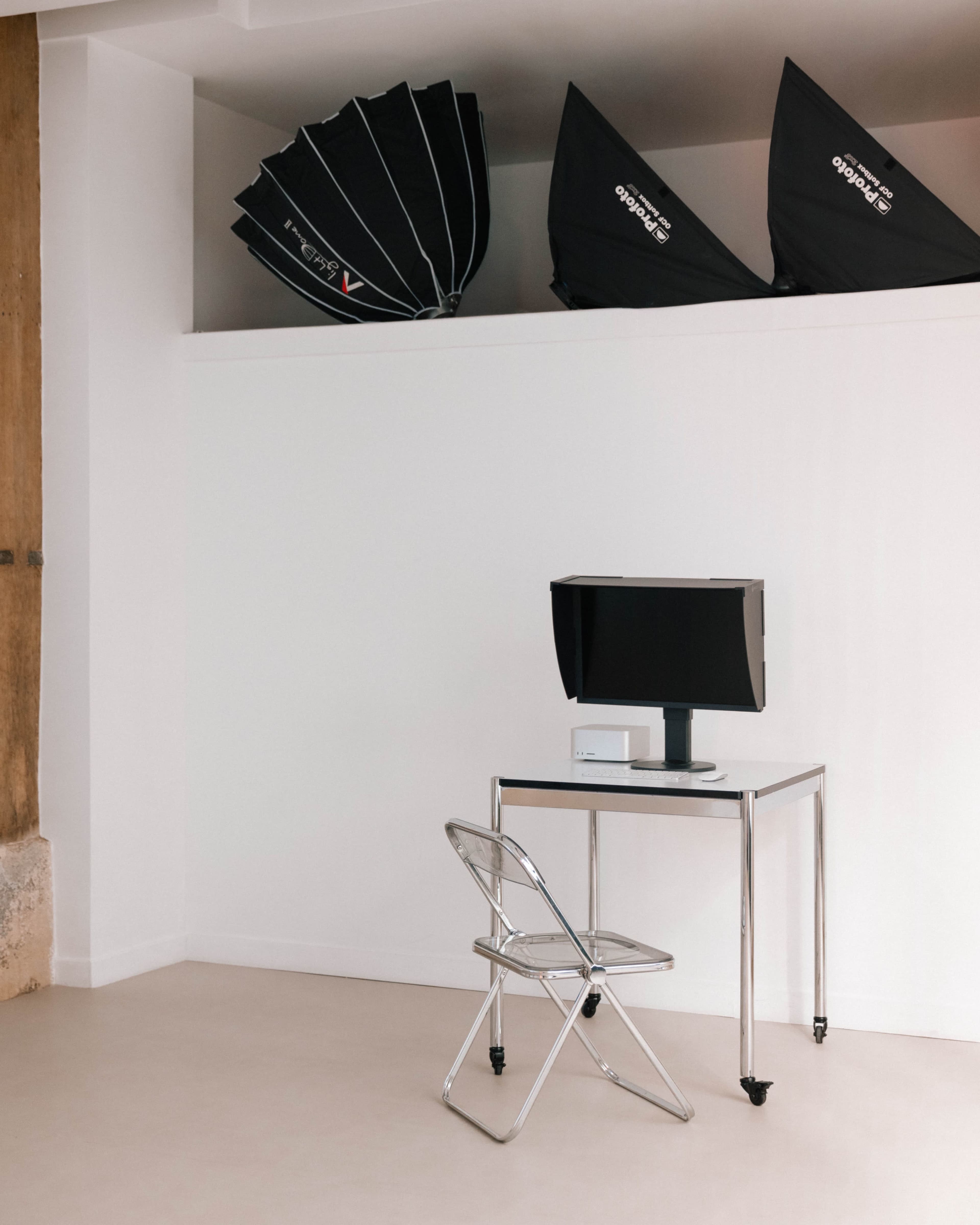 The image shows a minimalist workspace featuring a metal desk with a monitor and a folding chair, accompanied by several photography light umbrellas stored on a shelf above.
