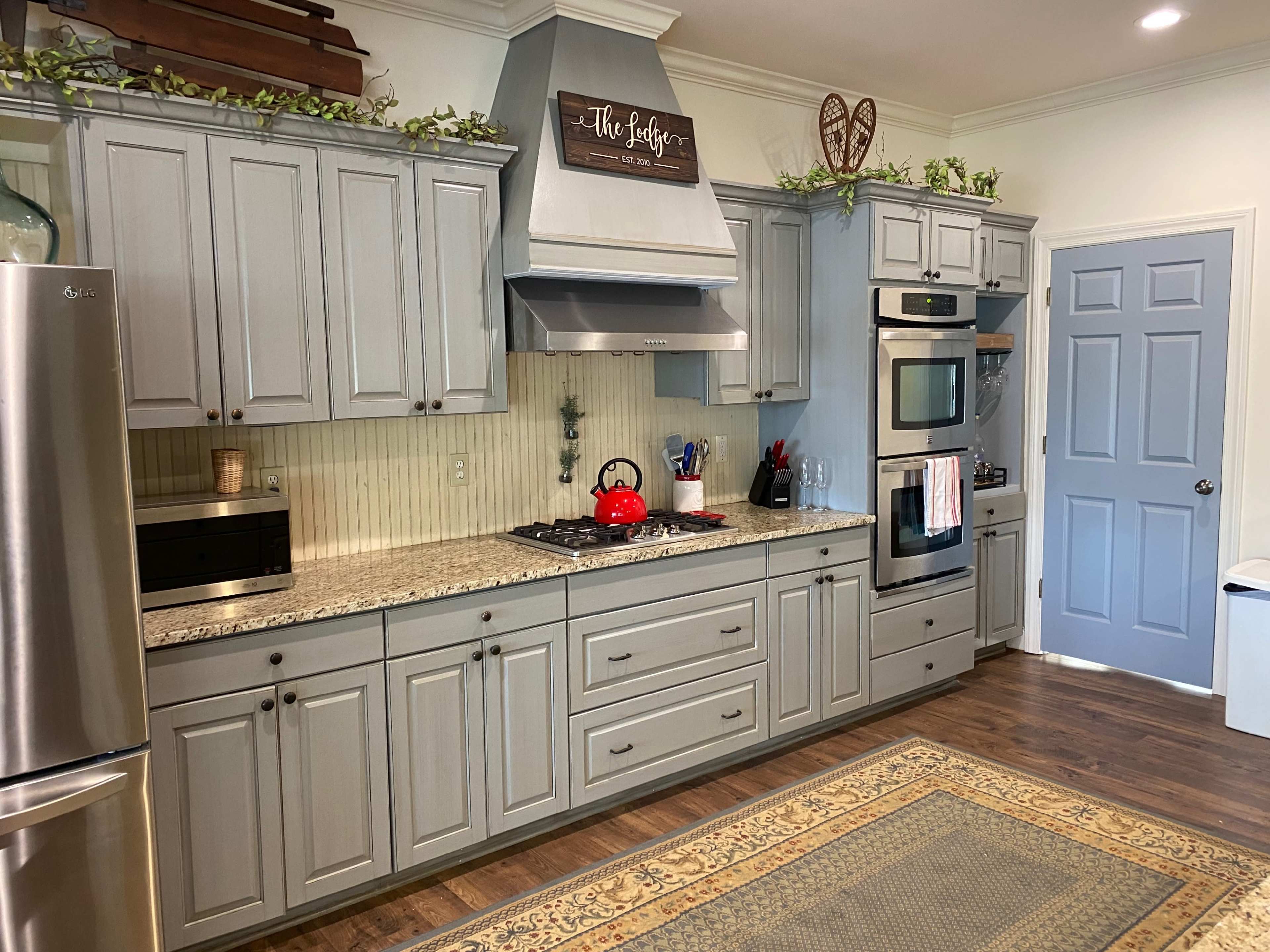 A modern kitchen with gray cabinets, a stainless steel refrigerator, a gas stove with a red kettle, and a decorative rug on the wooden floor.