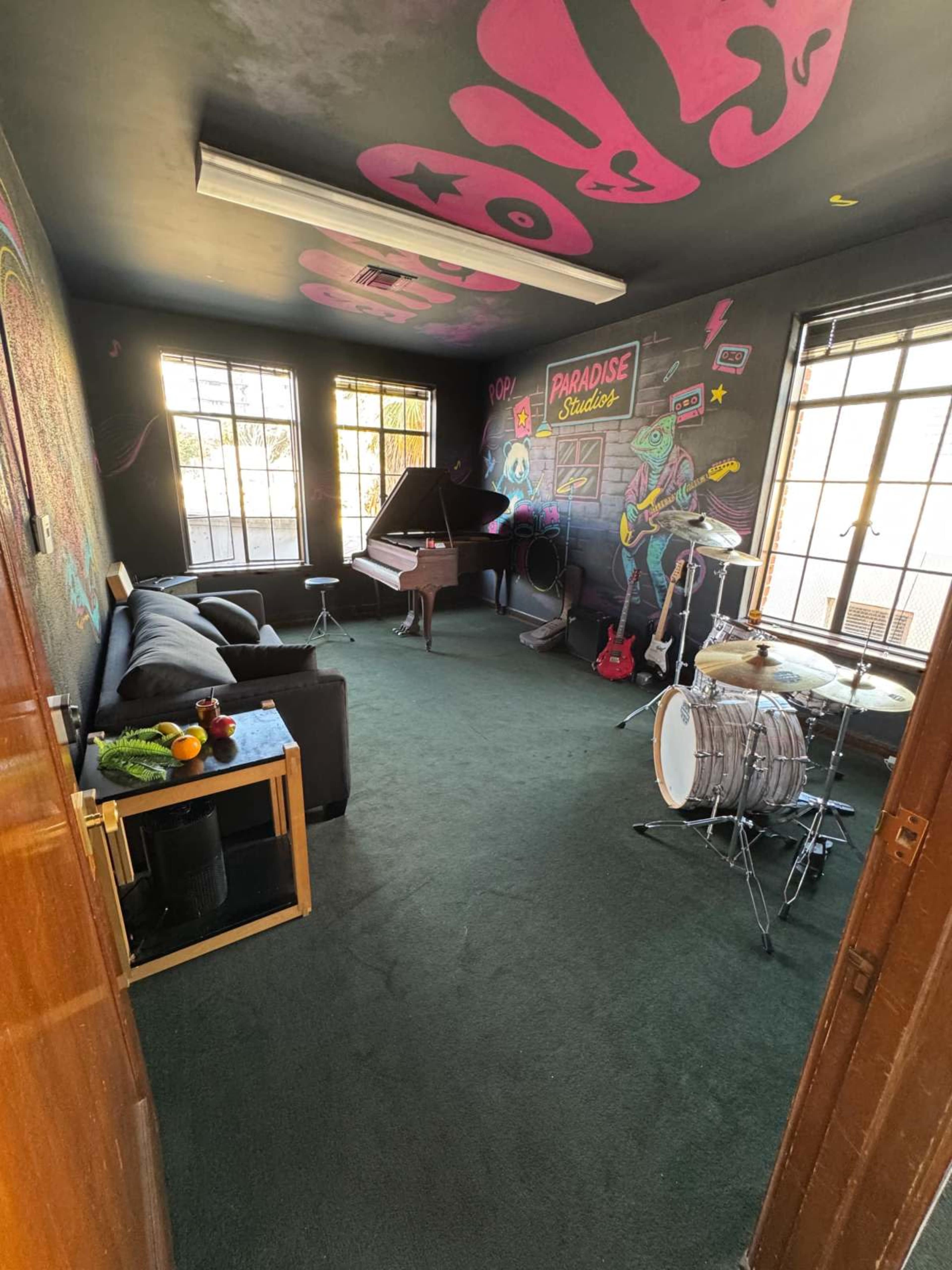 The image shows a music studio featuring a grand piano, a drum set, a sofa, a table with fruits, and colorful wall artwork.