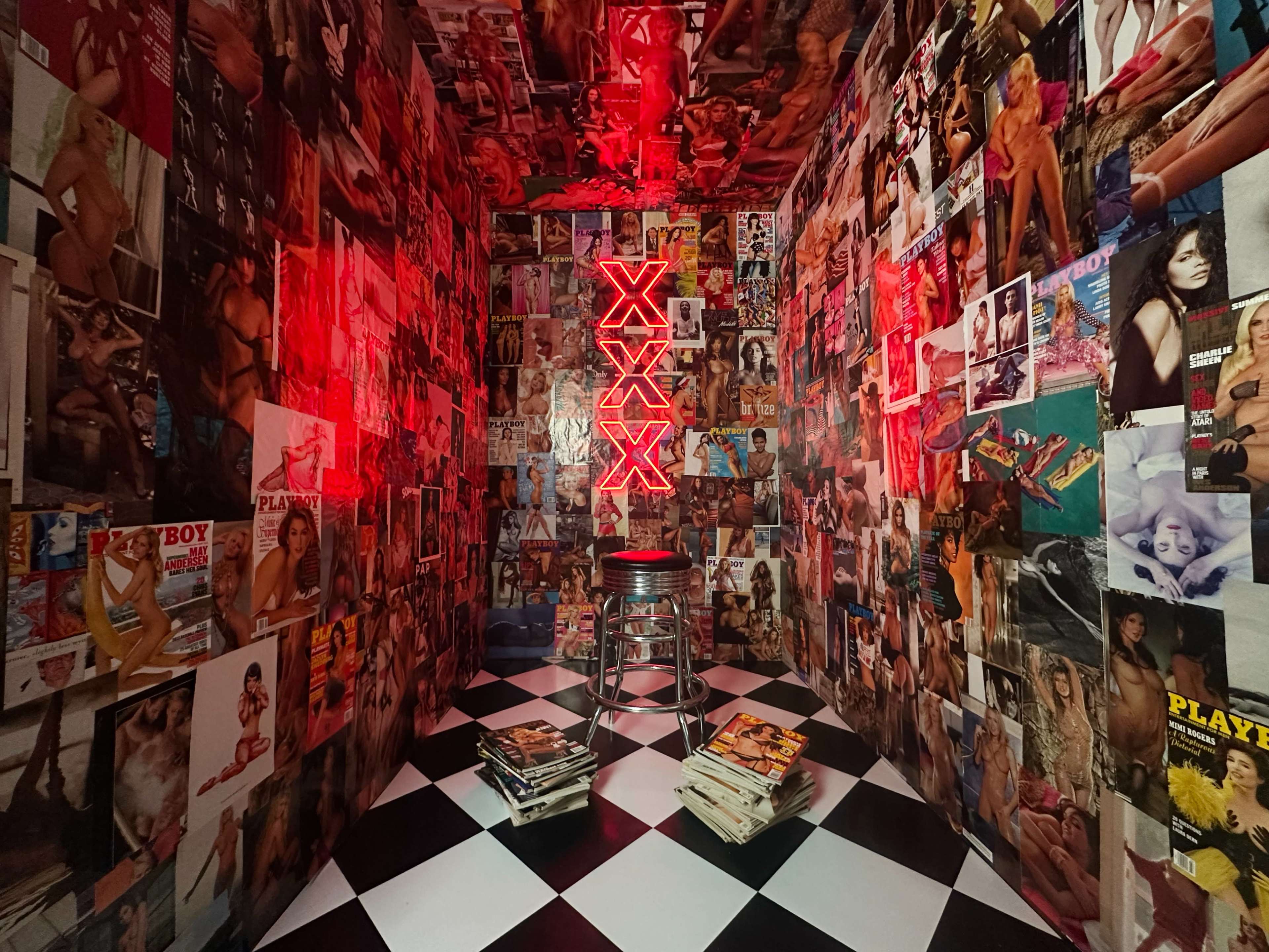 The image shows a small room covered with posters and magazine clippings, featuring a neon sign that reads "XXXX" in red, and a stool in the center on a black and white checkered floor.