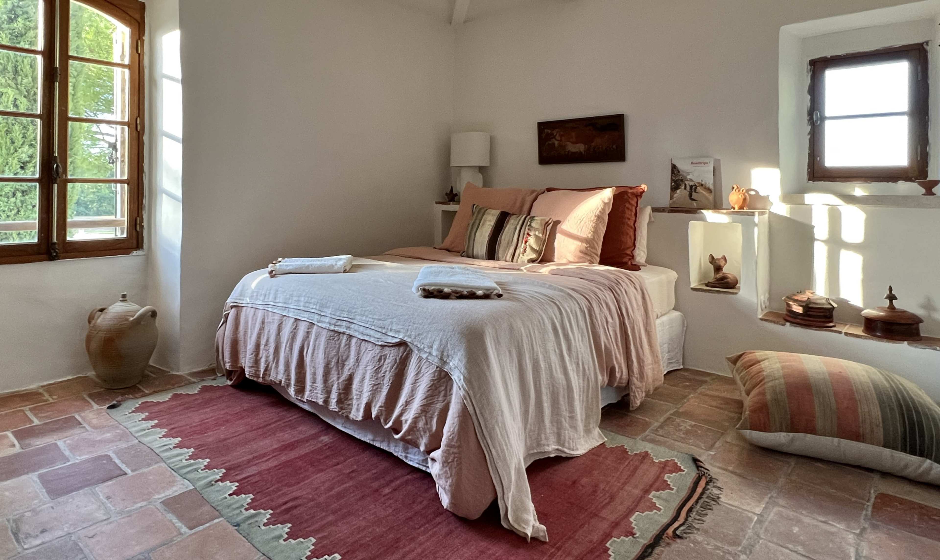 A cozy bedroom features a bed with light-colored linens, a patterned rug, and windows allowing natural light to filter in.