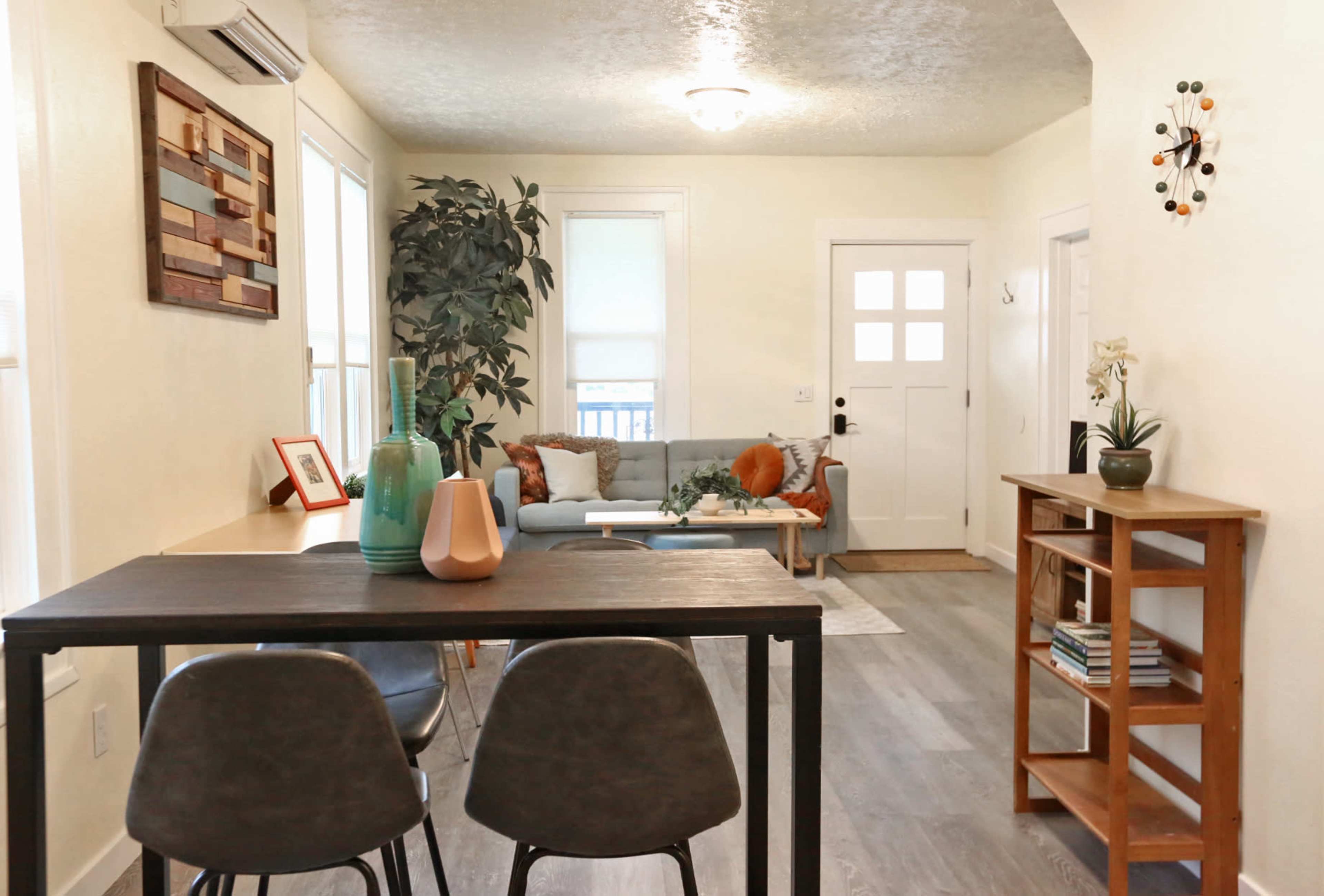 A cozy living area features a dining table with chairs, a blue sofa, a wooden shelf, and a plant near a front door with a window.