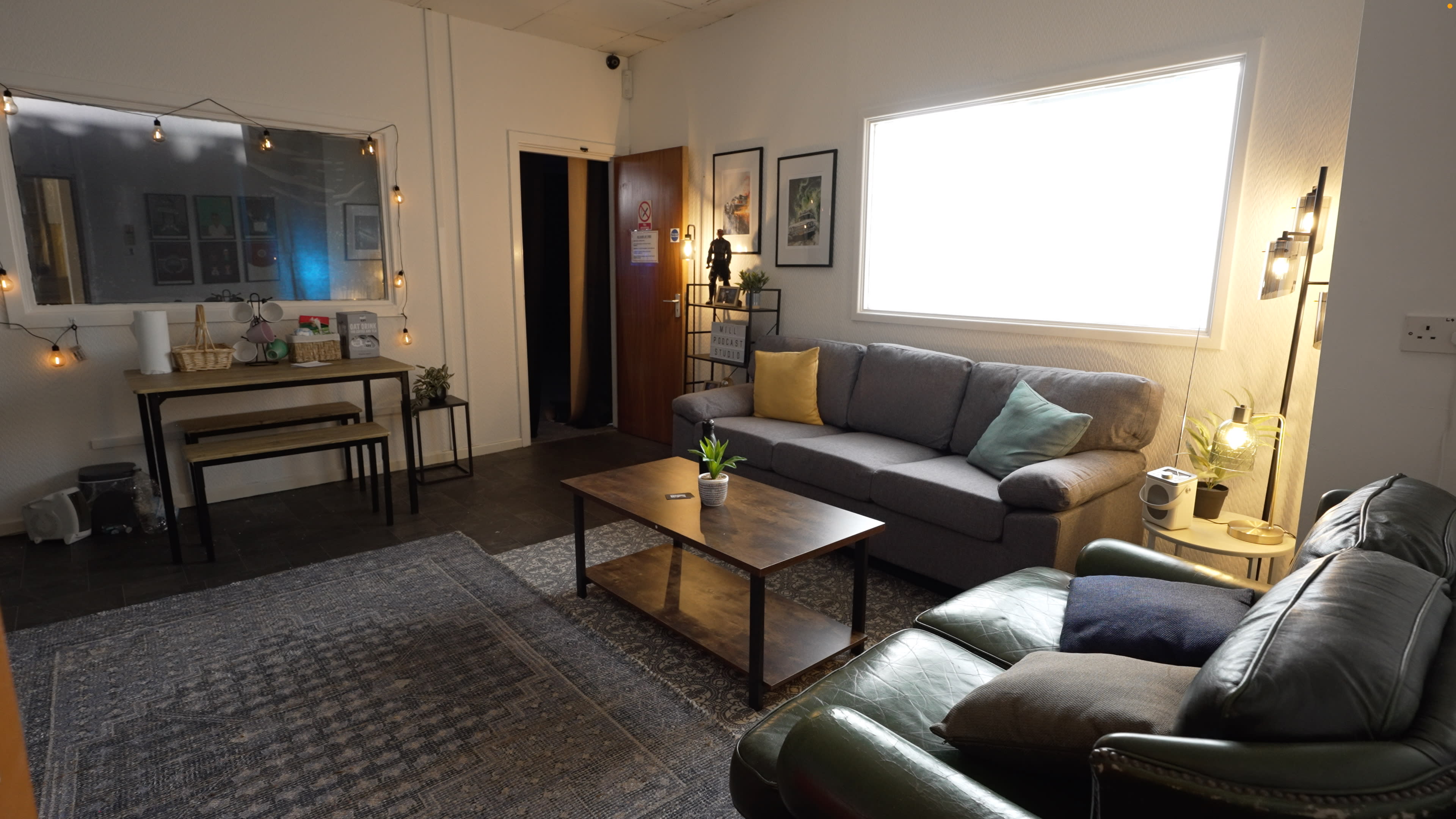 The image shows a cozy living room with a gray sofa, a green leather couch, a wooden coffee table, and a small dining table, all illuminated by string lights and natural light from a window.