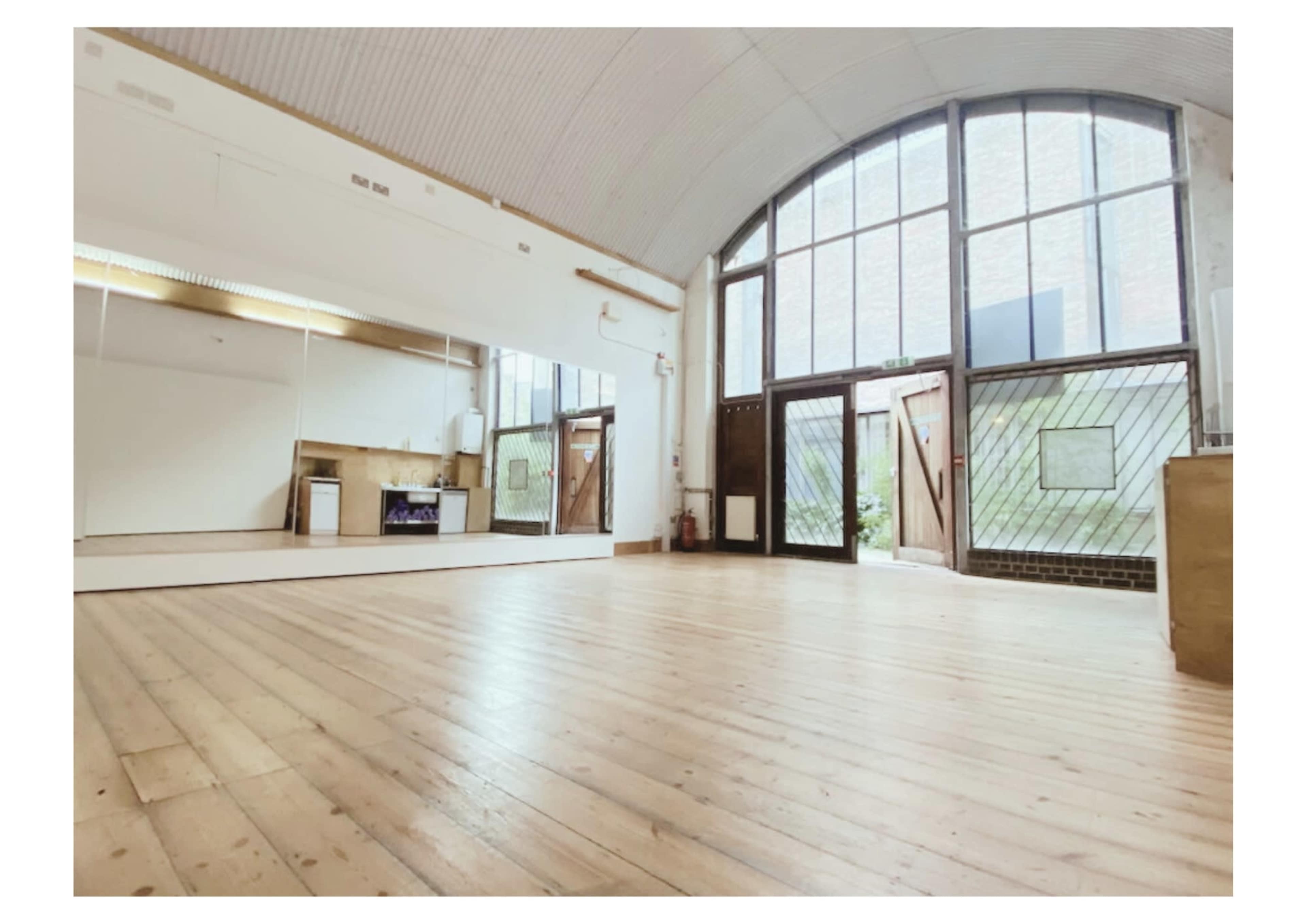 A spacious room with wooden flooring, large windows, and mirrors on one wall, leading to an outdoor area.