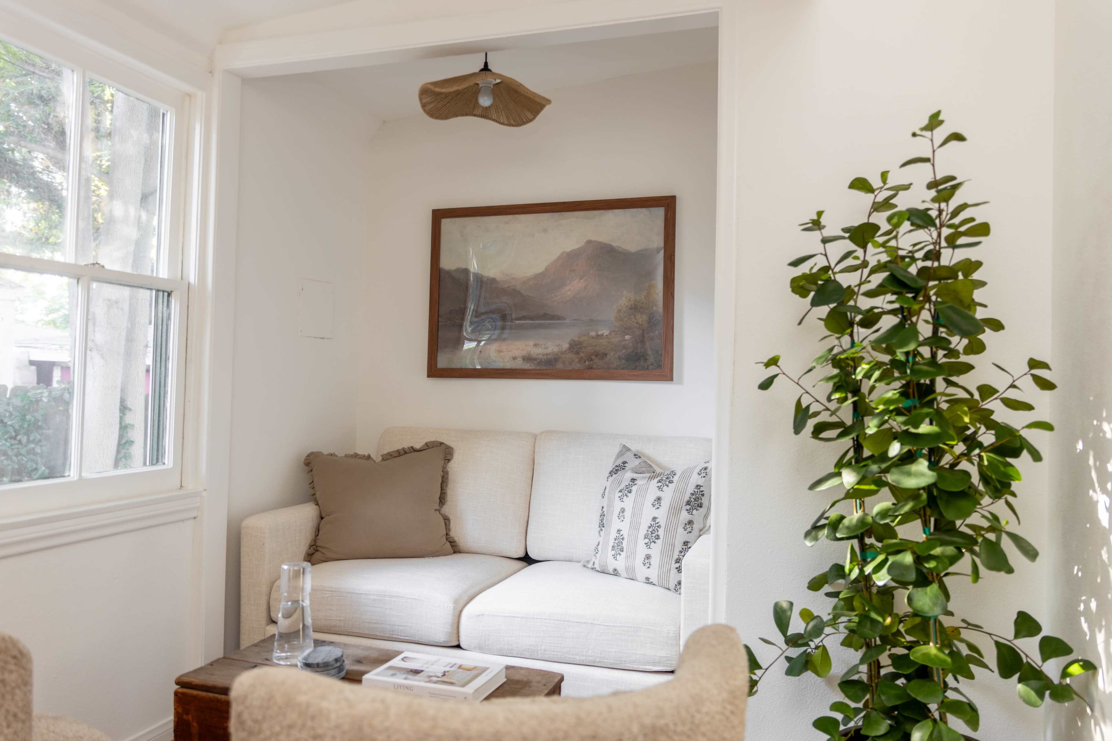 The image shows a small, cozy corner of a room with a white couch, a framed landscape painting on the wall, and a potted plant nearby.