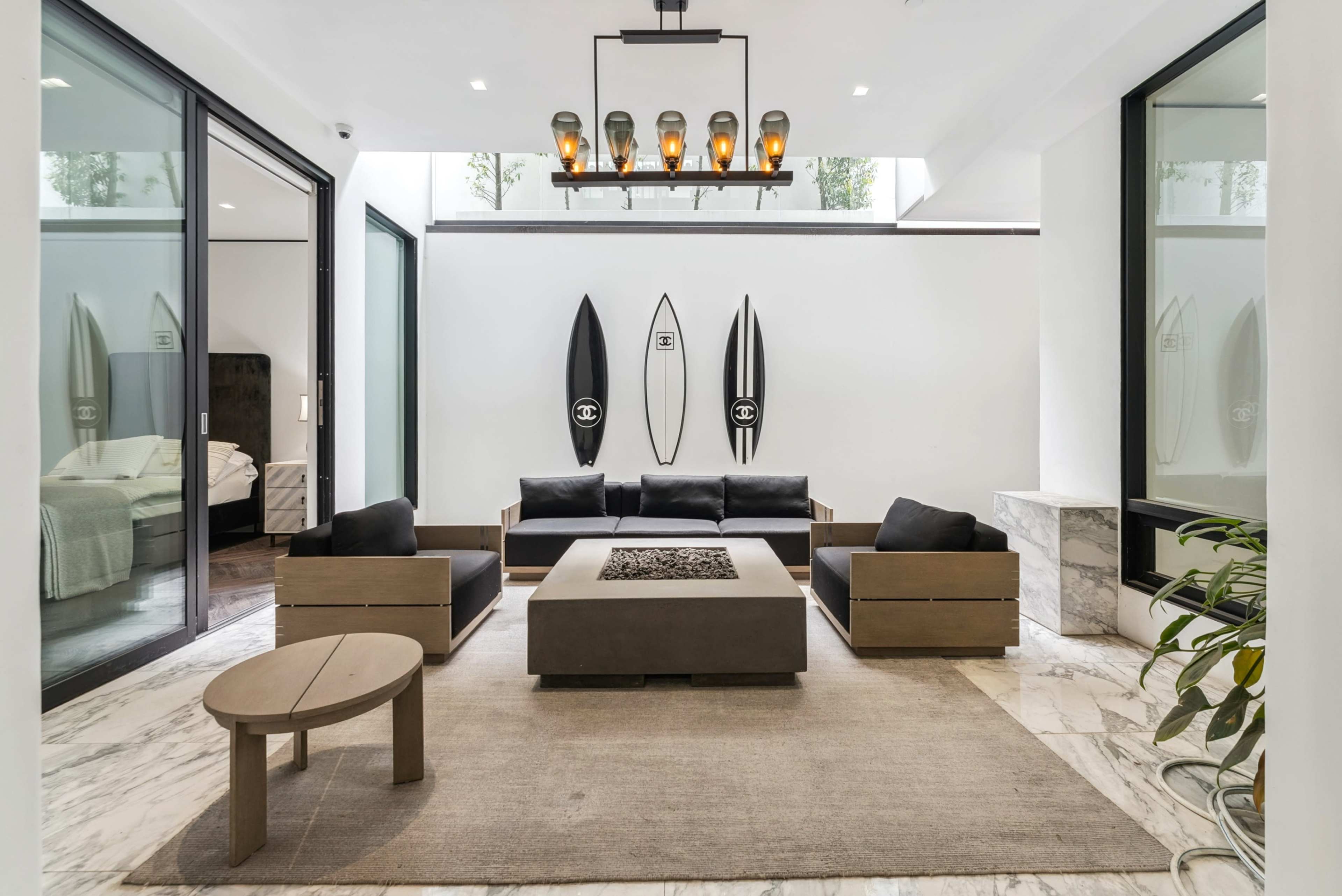 The image shows a modern living room with a central coffee table, black sofas, a light rug, and surfboards mounted on the wall.