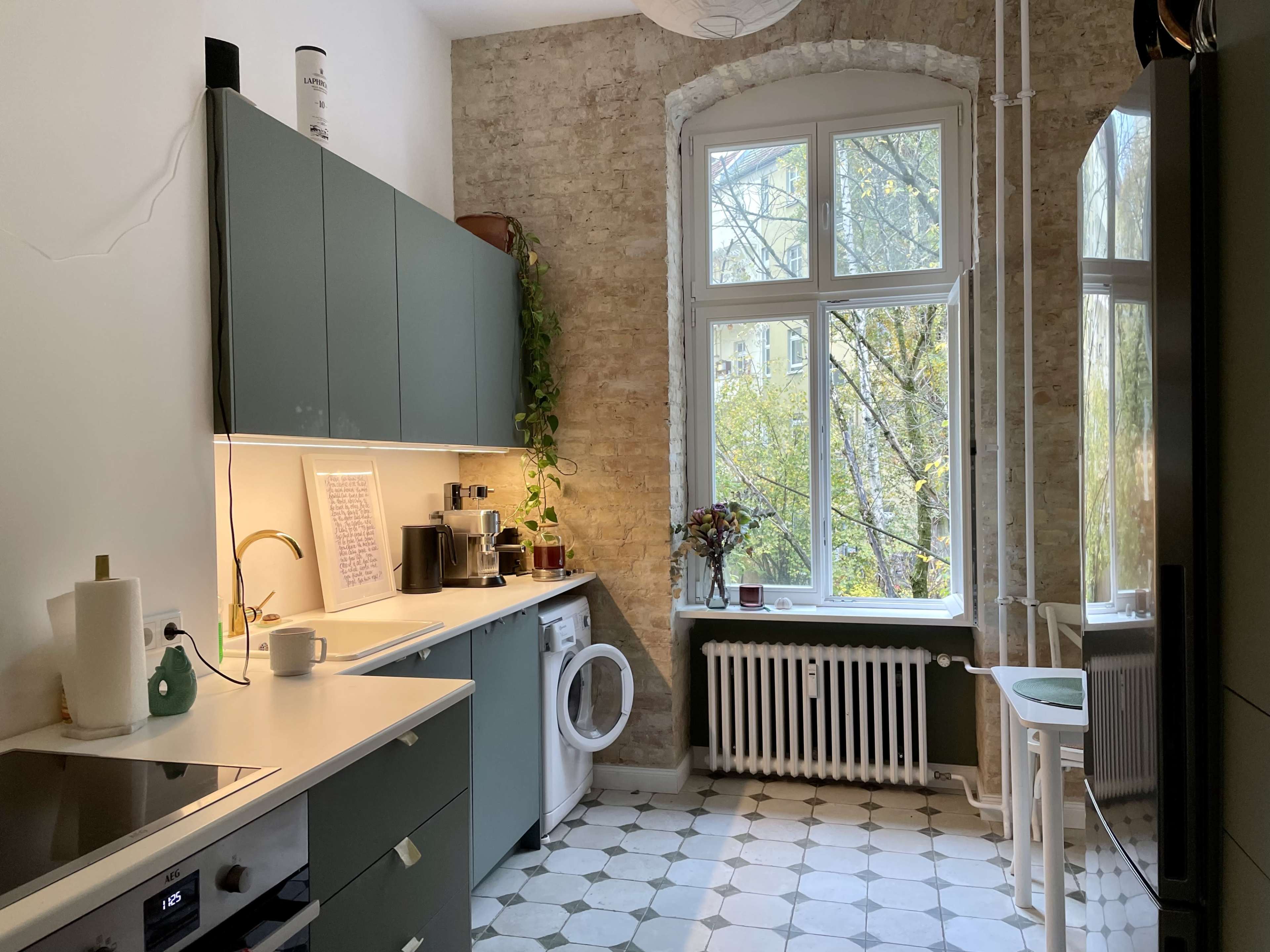 A modern kitchen features green cabinetry, a washing machine, and a large window overlooking trees.