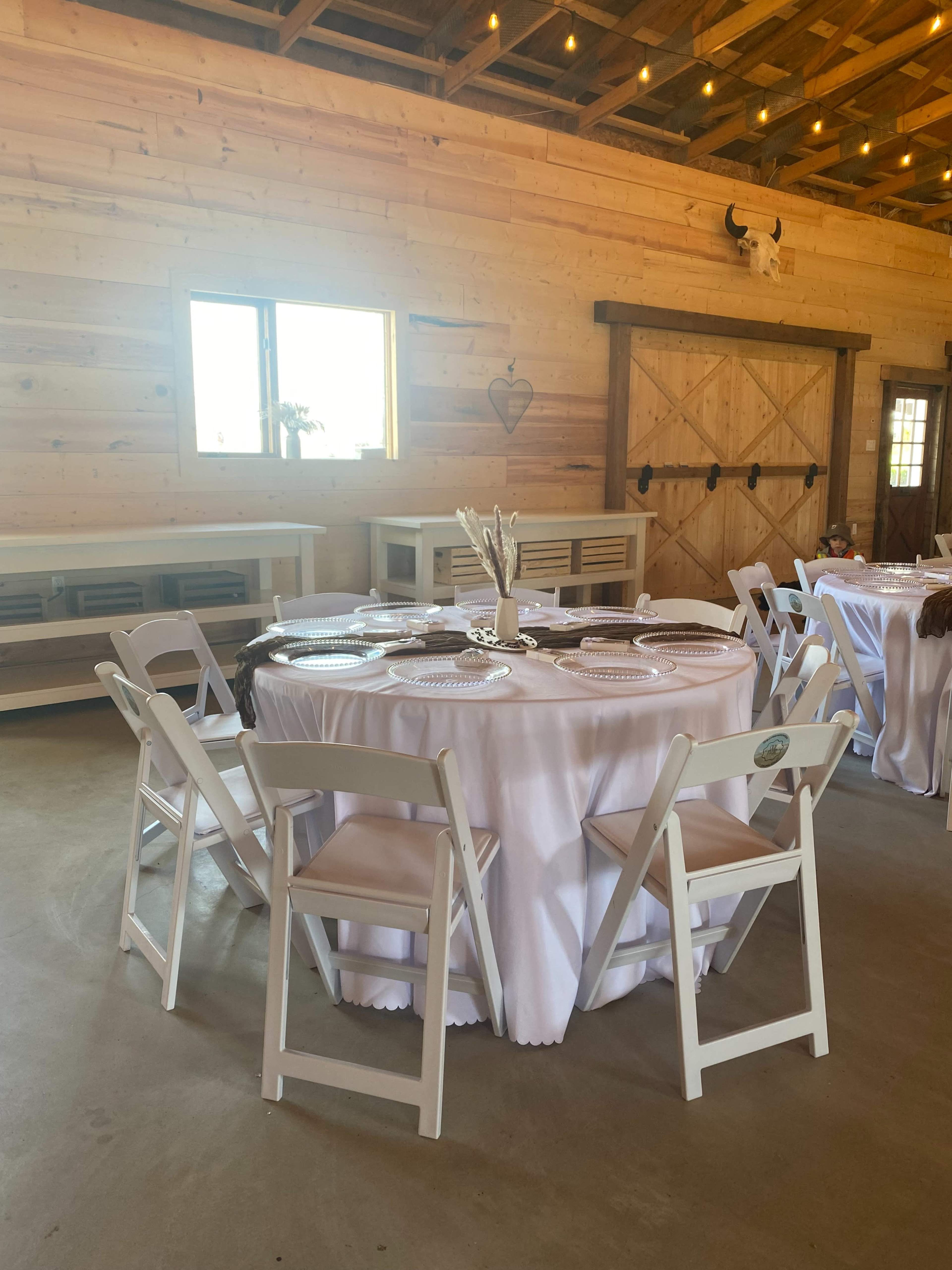 A round table set with white tablecloth and plates, surrounded by white folding chairs in a wooden interior space.