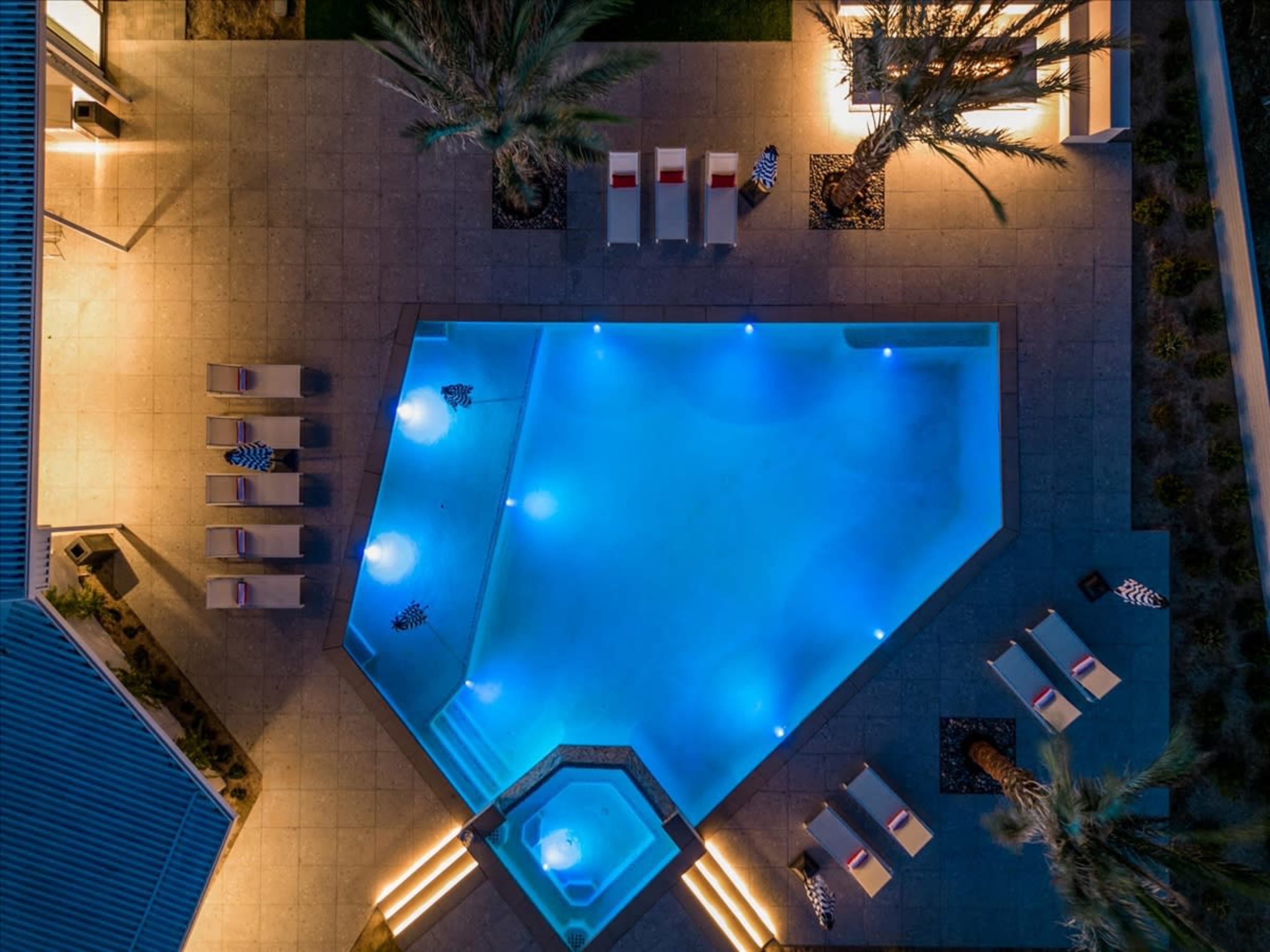 An aerial view of a geometric swimming pool surrounded by lounge chairs and illuminated pathways at night.