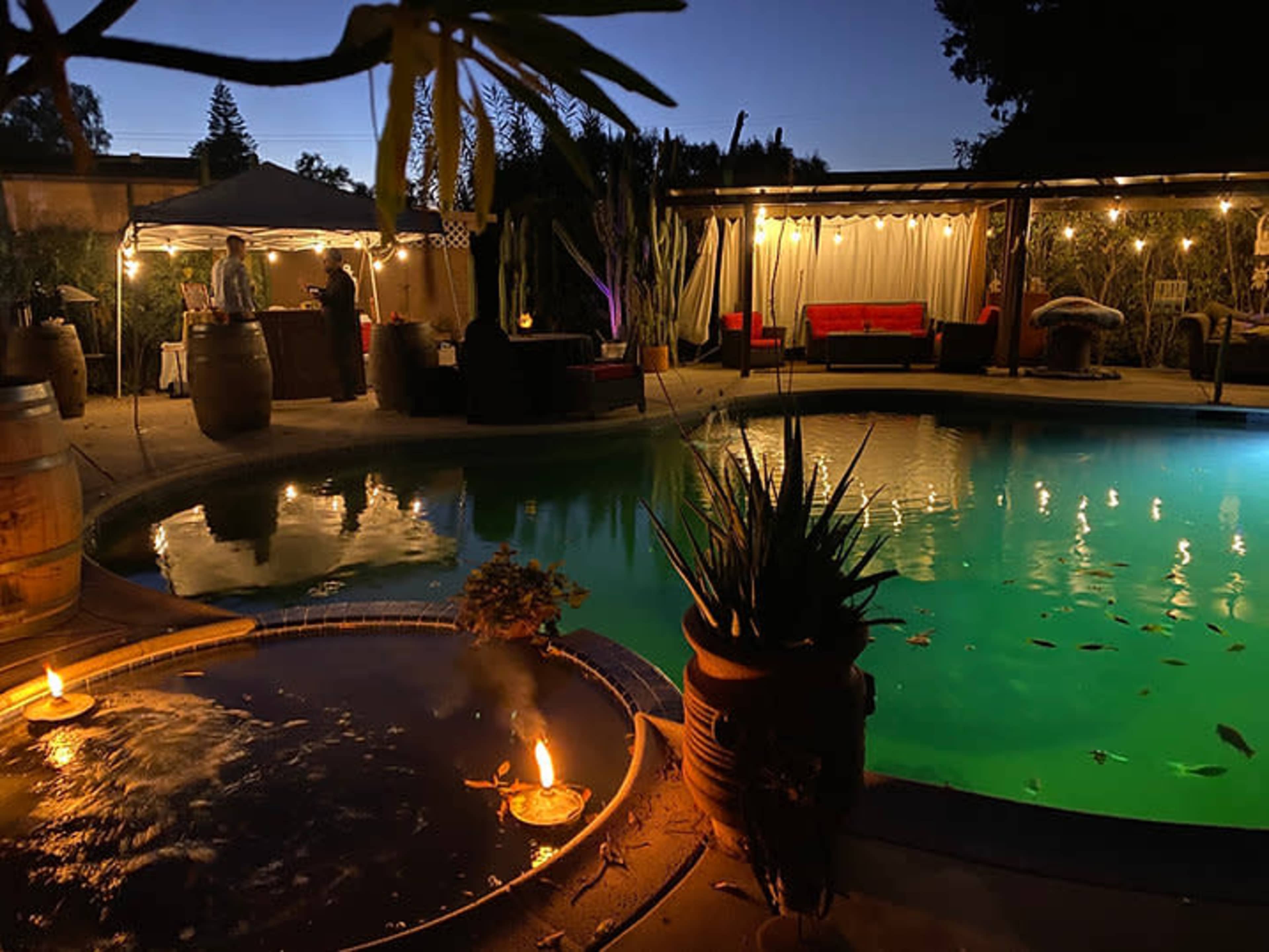 A pool area at night features a lit water feature beside the pool, with surrounding greenery and a lounge area illuminated by soft lighting.