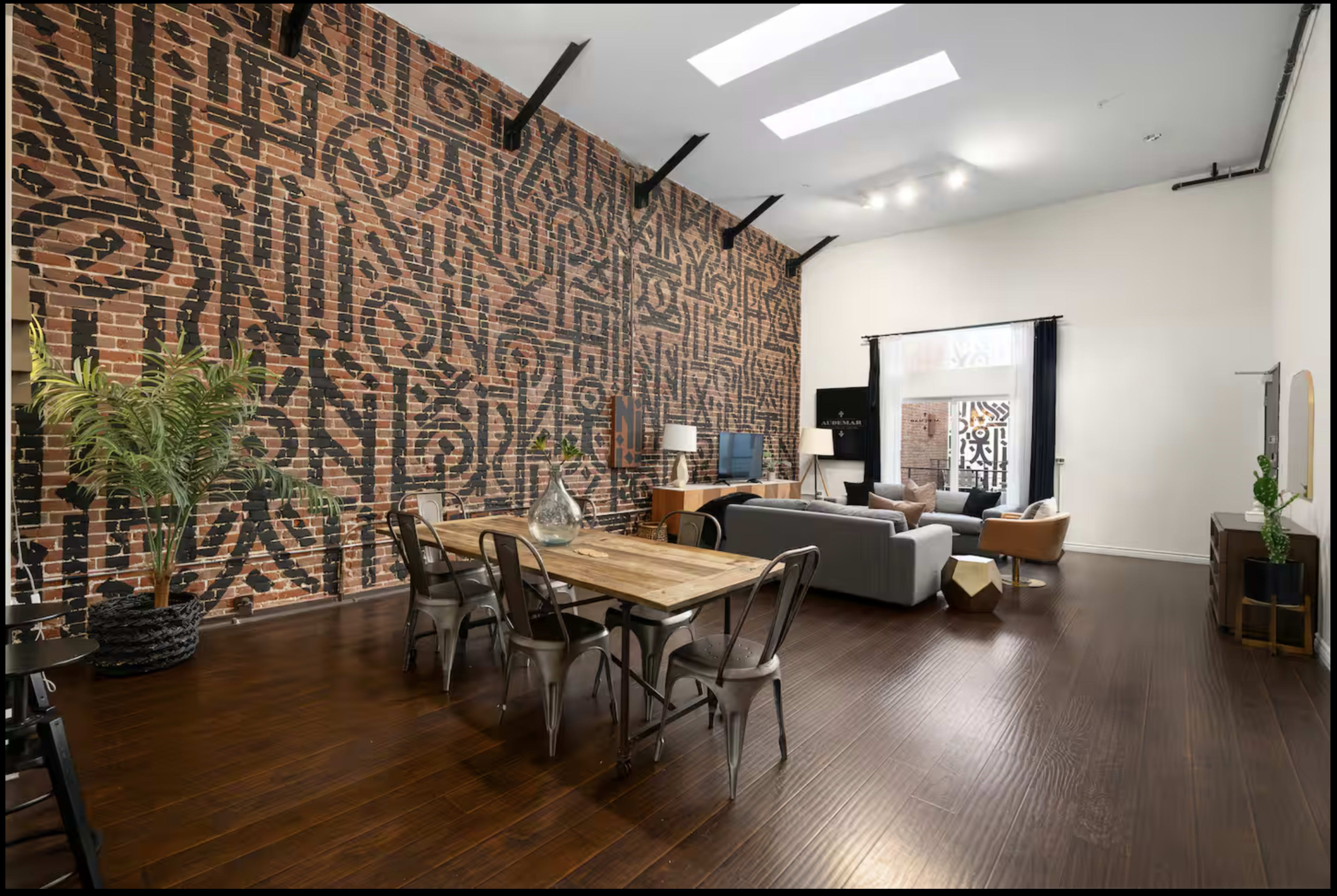 The image shows a spacious living area featuring a brick wall with abstract designs, a wooden dining table, a sofa, and large windows allowing natural light to enter.
