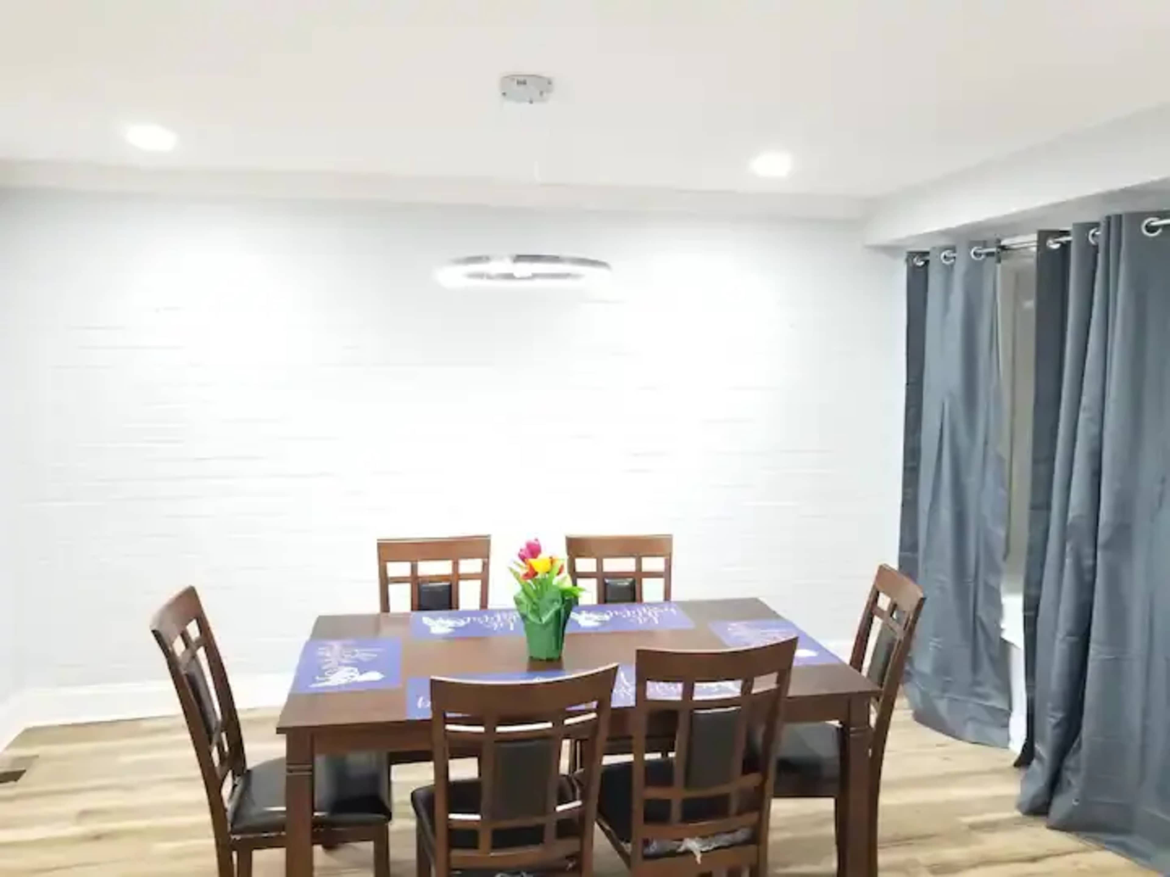 A wooden dining table surrounded by six chairs is set against a light-colored wall, with a bouquet of flowers in the center and curtains on the windows.
