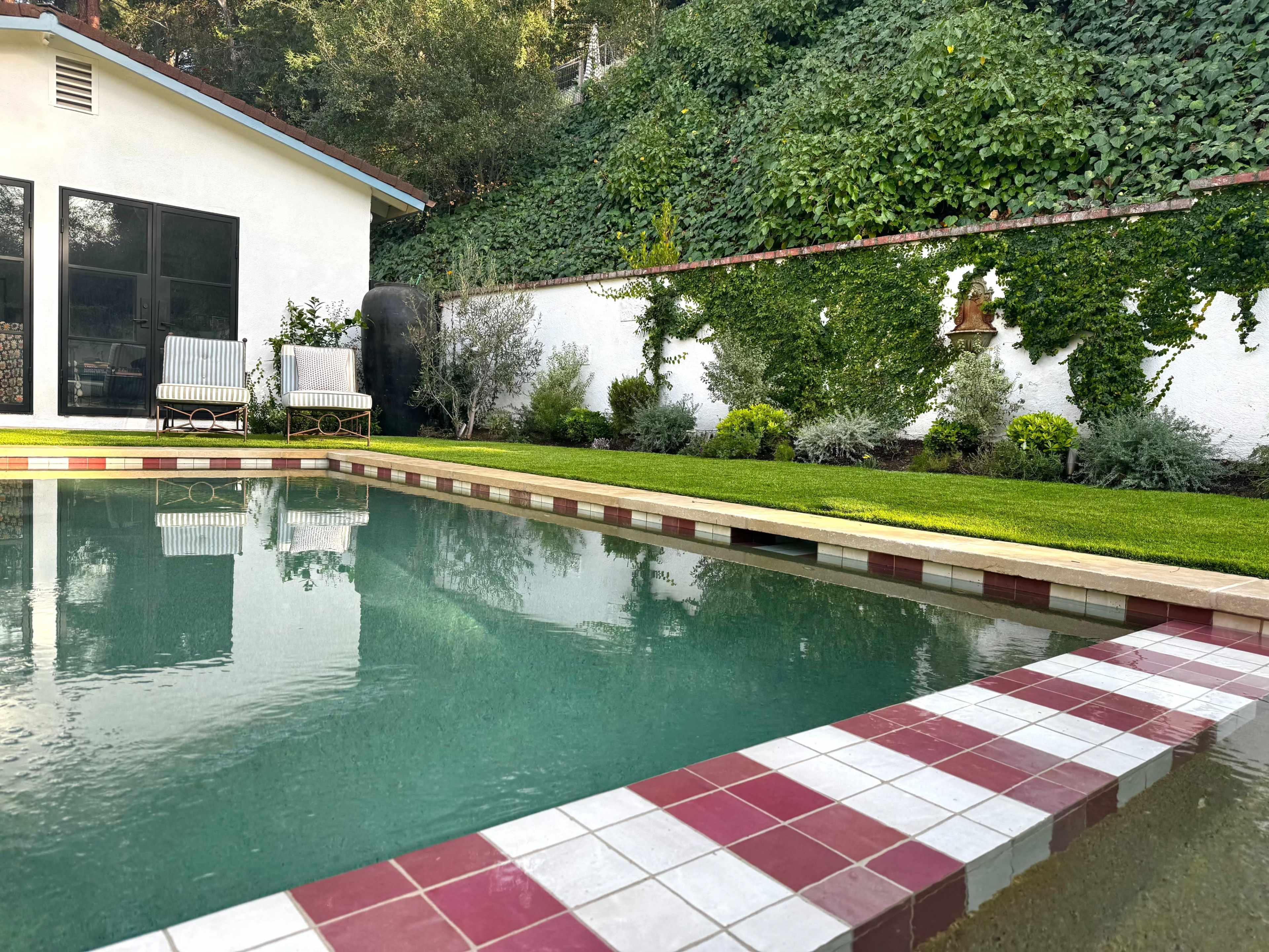 A well-maintained swimming pool is bordered by a tiled edge and surrounded by a lush green lawn and bushes.