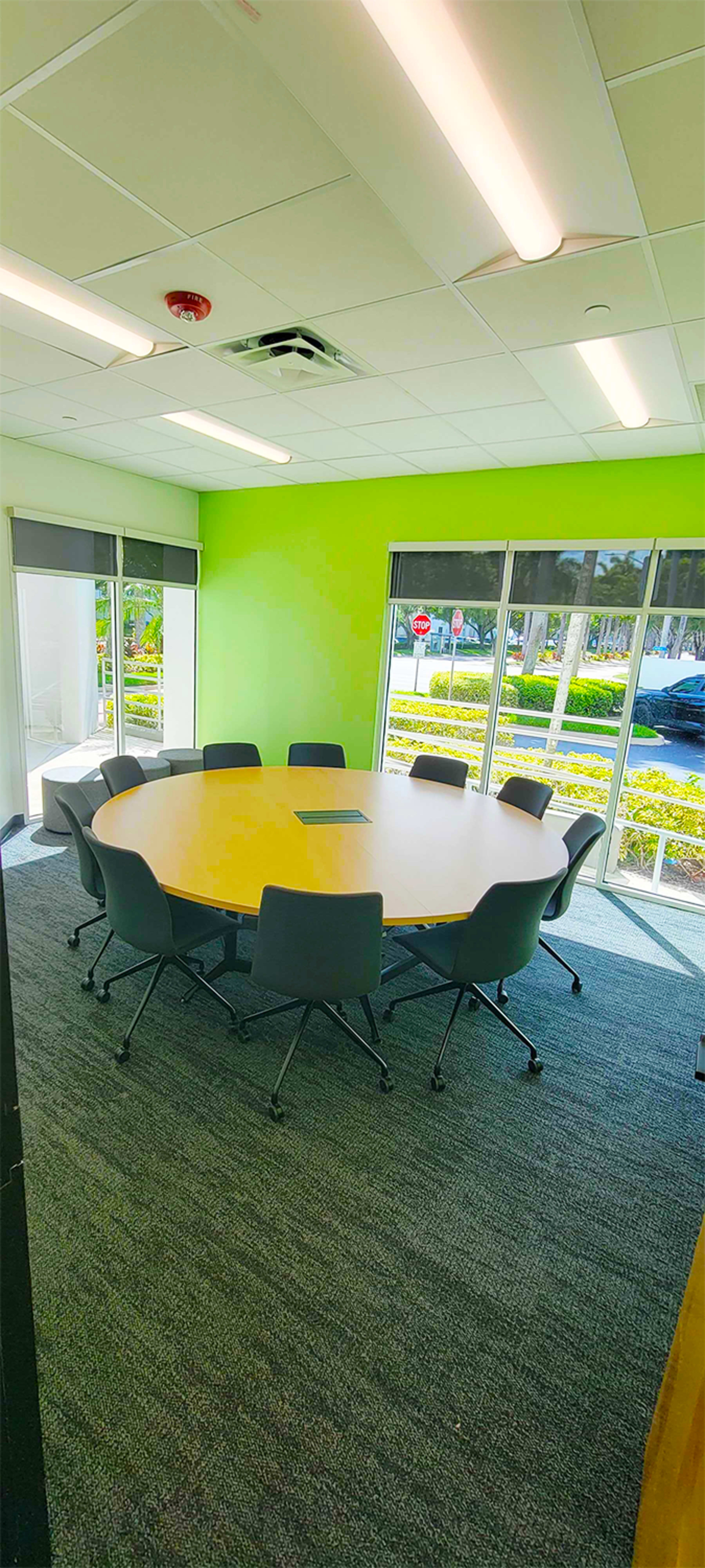 A conference room with a round table and ten chairs is illuminated by natural light from large windows, featuring a vibrant green wall.