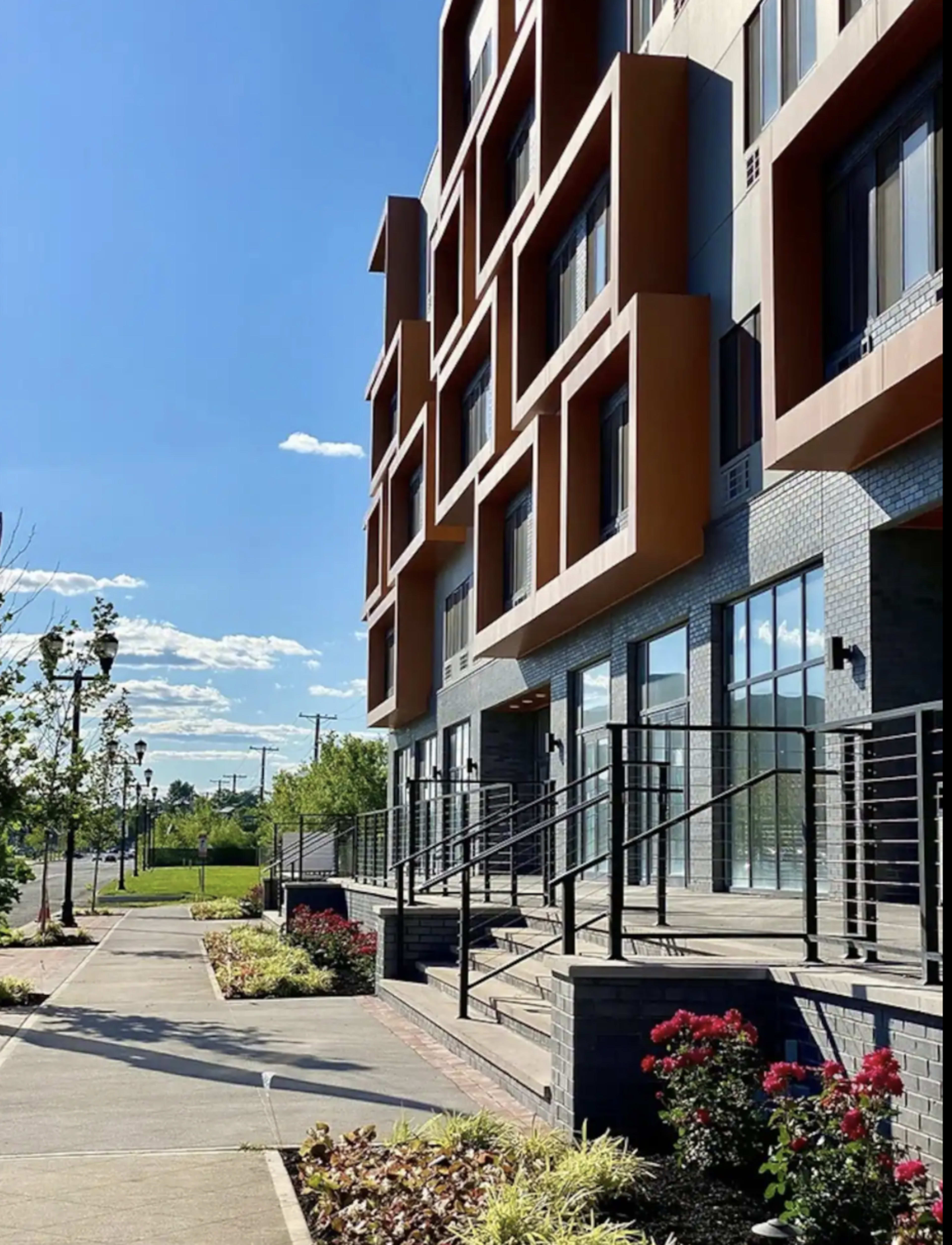 The image shows a modern building with angular architectural features beside a paved walkway lined with flowers and greenery.
