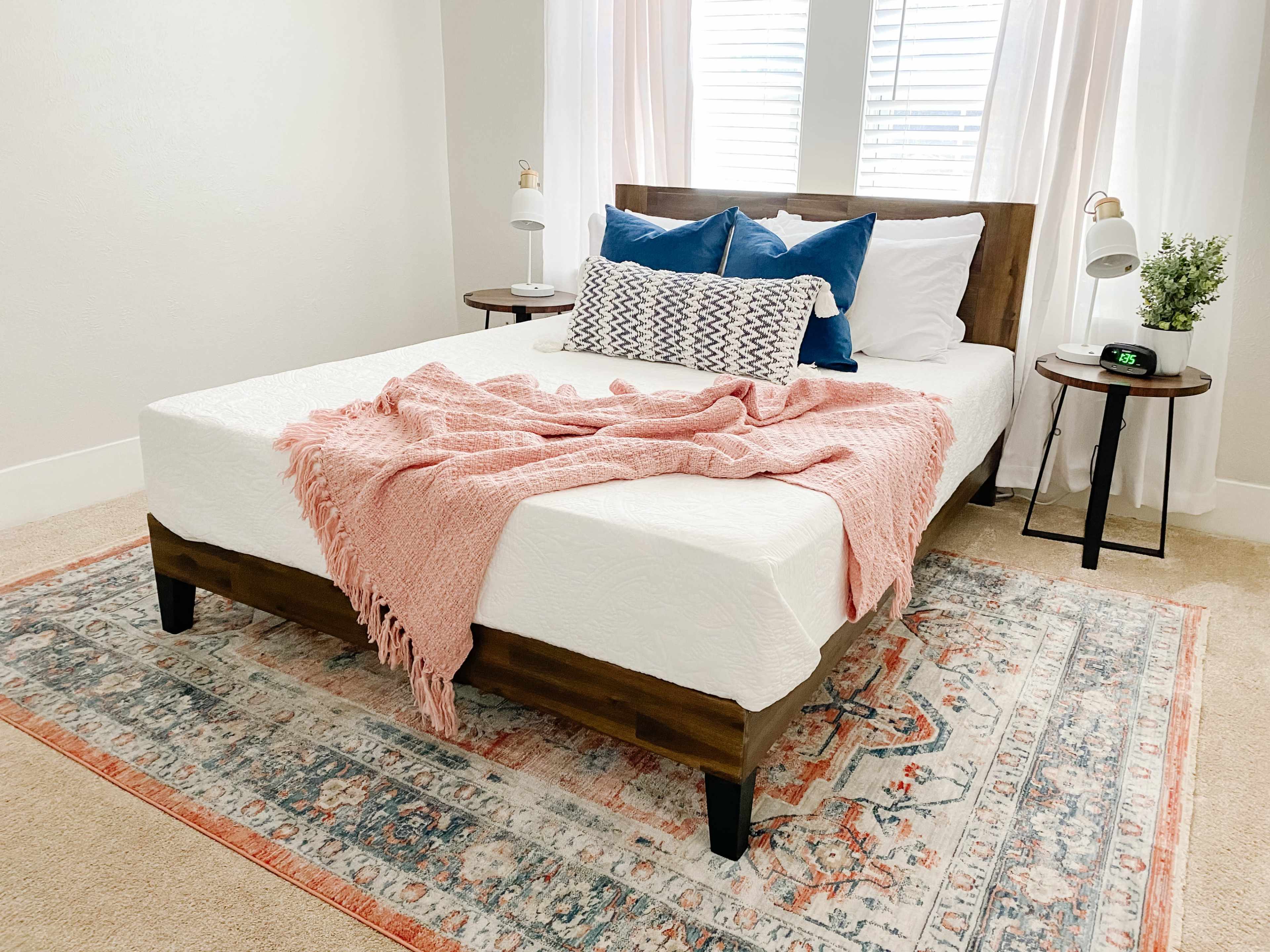 A bed with a light pink throw blanket and decorative pillows is positioned between two nightstands on a patterned area rug in a softly lit room.