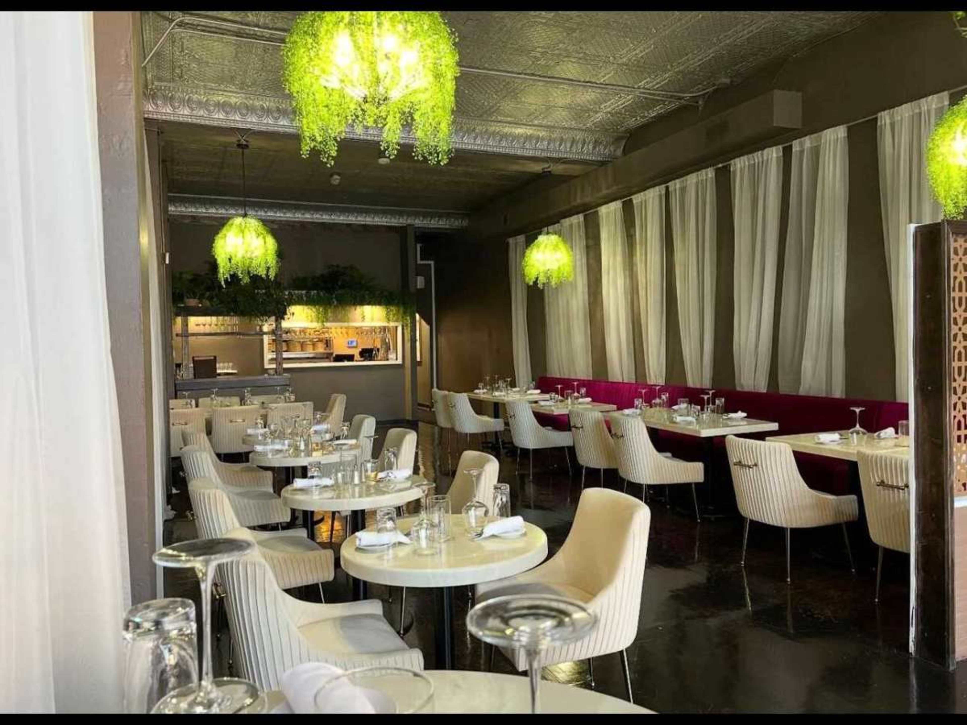 The image shows an elegant restaurant interior with white tables and chairs, green hanging lights, and soft curtains along the windows.
