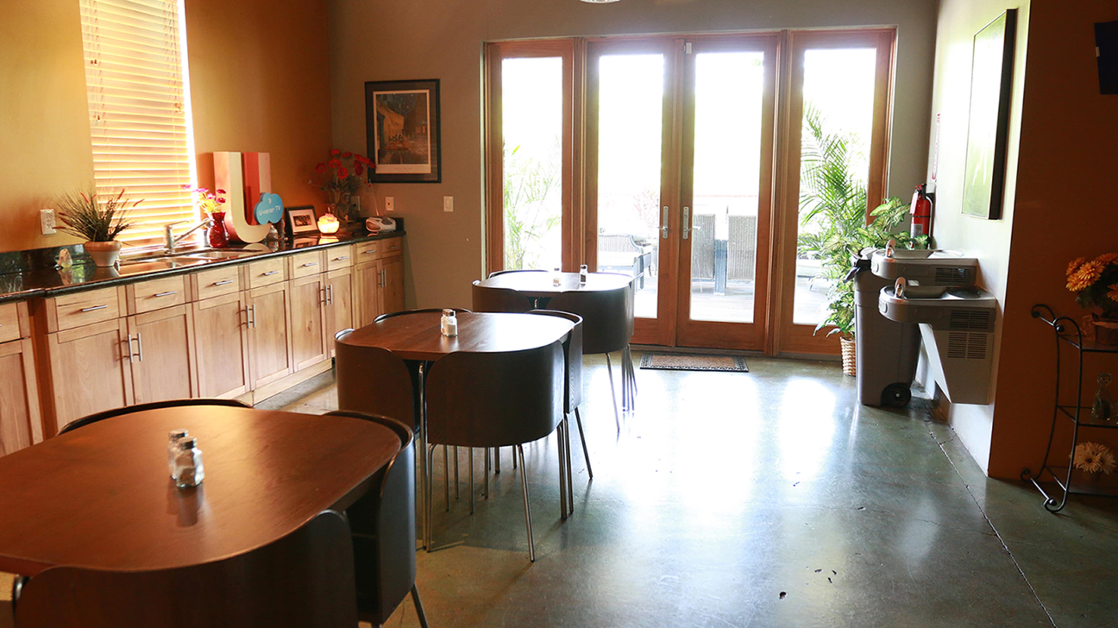 The image shows a small café with wooden tables, a countertop area, and large doors leading to an outdoor space.
