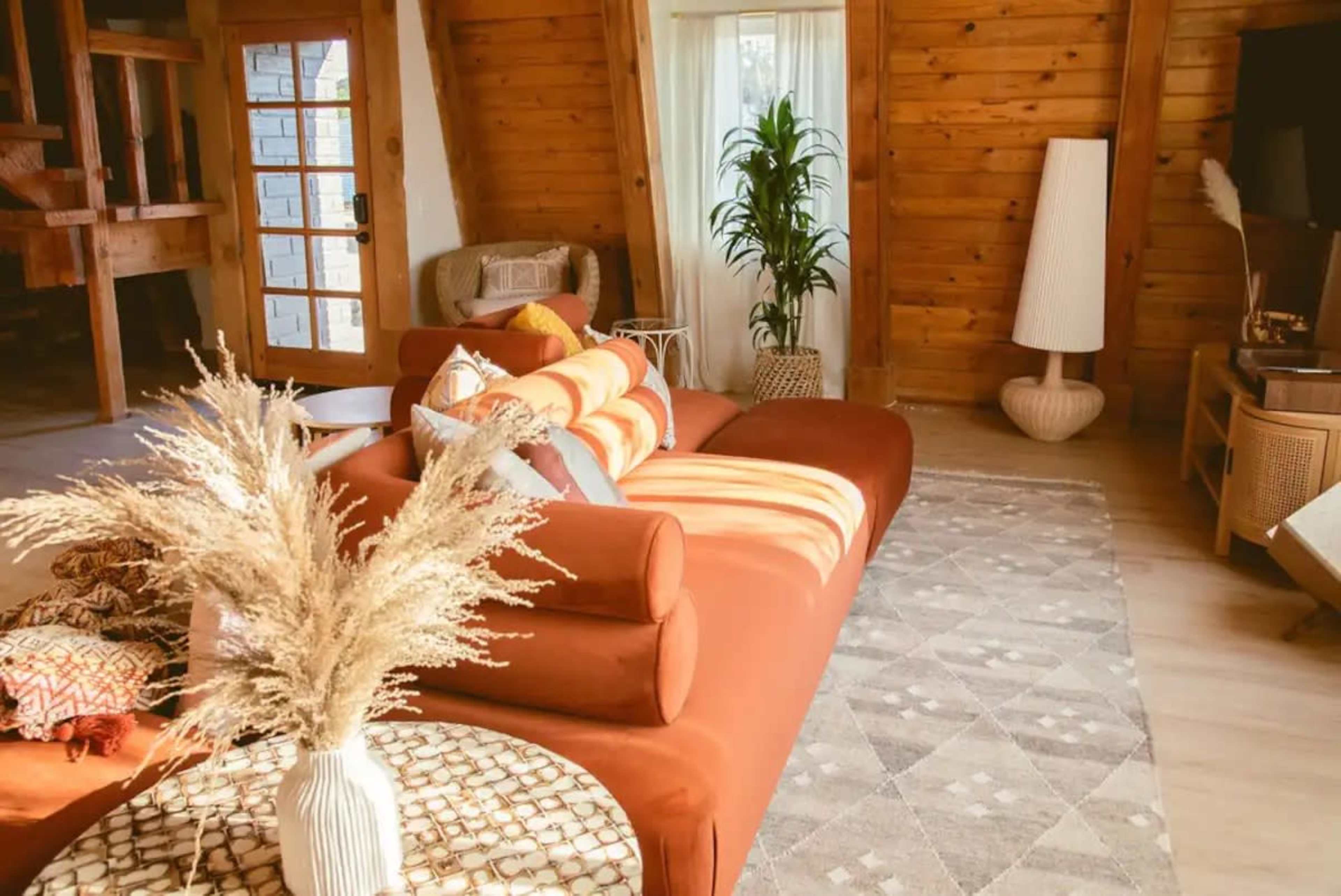 The scene features a cozy living room with a large orange sectional sofa, a round patterned coffee table, and a mix of plants and decorative elements in a wooden-paneled space.