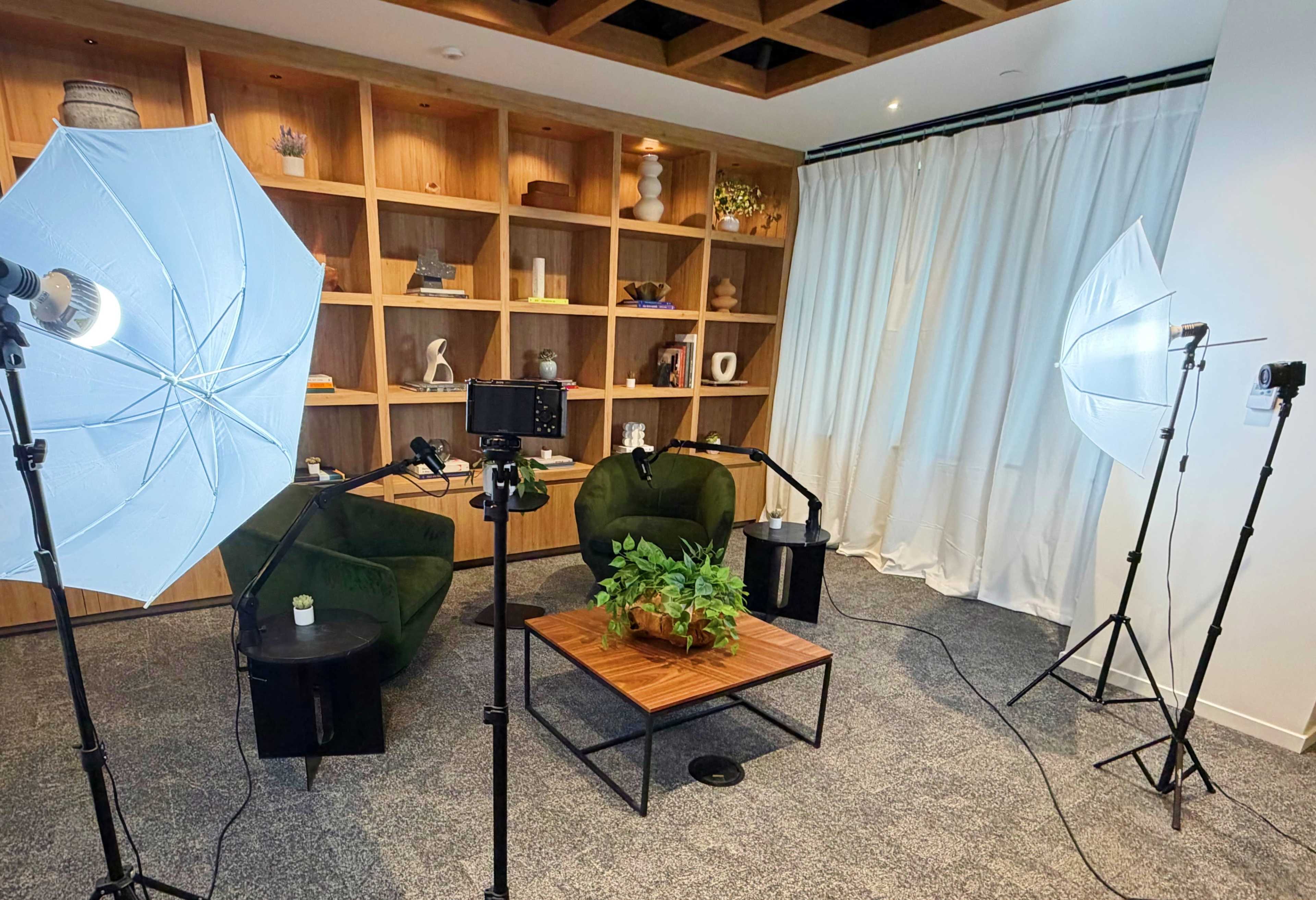 The image shows a small studio setup featuring two green chairs, a wooden coffee table with a plant, and two softbox lights positioned on tripods, with a backdrop of a wooden shelf filled with decorative items.