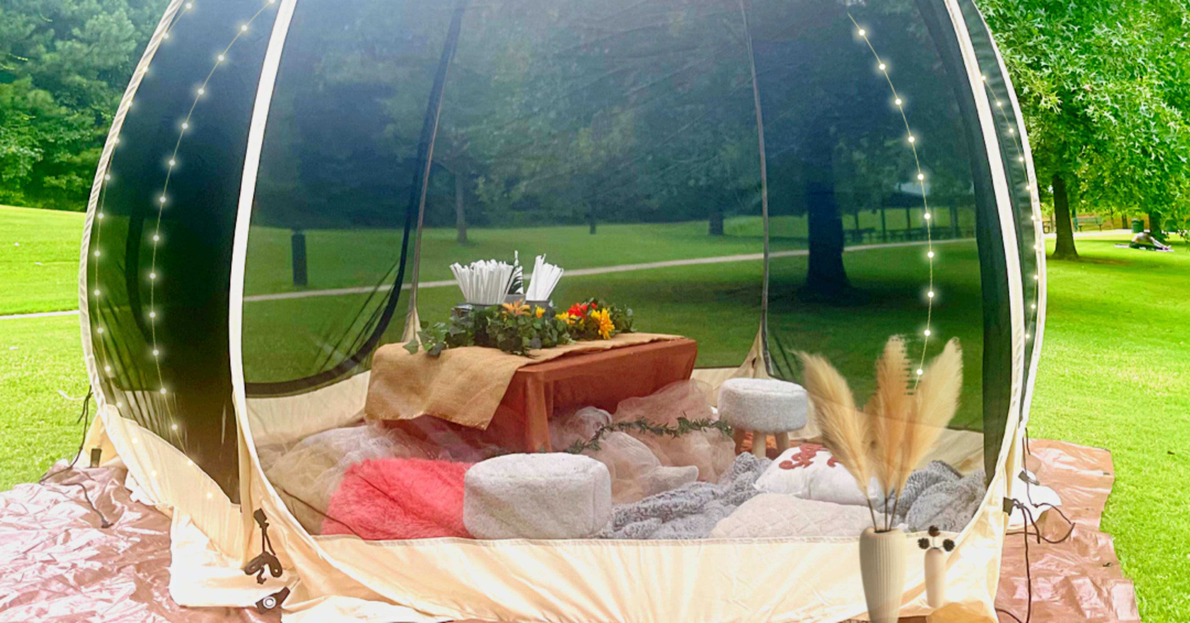 A transparent dome tent is set up on a grassy area, furnished with cushions, blankets, and a small table adorned with decorative items.