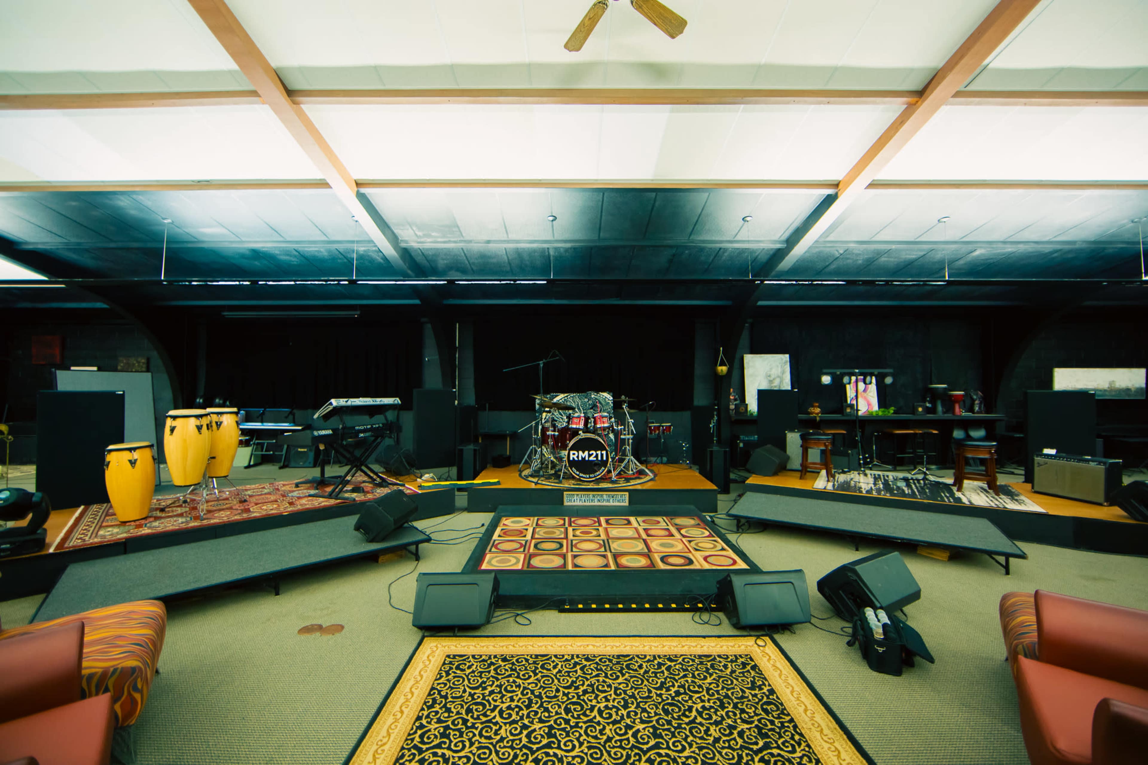 The image shows a spacious music studio equipped with instruments, including drums, keyboards, and conga drums, arranged on a stage with sound equipment in the background.