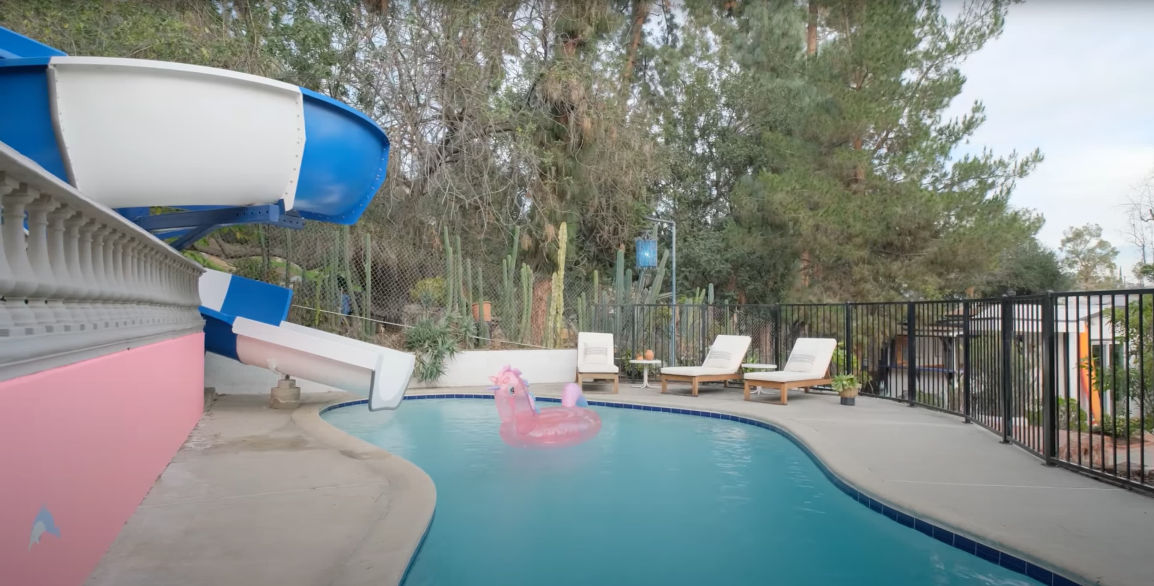 A swimming pool features a blue water slide, a pink flamingo float, and three lounge chairs beside it.