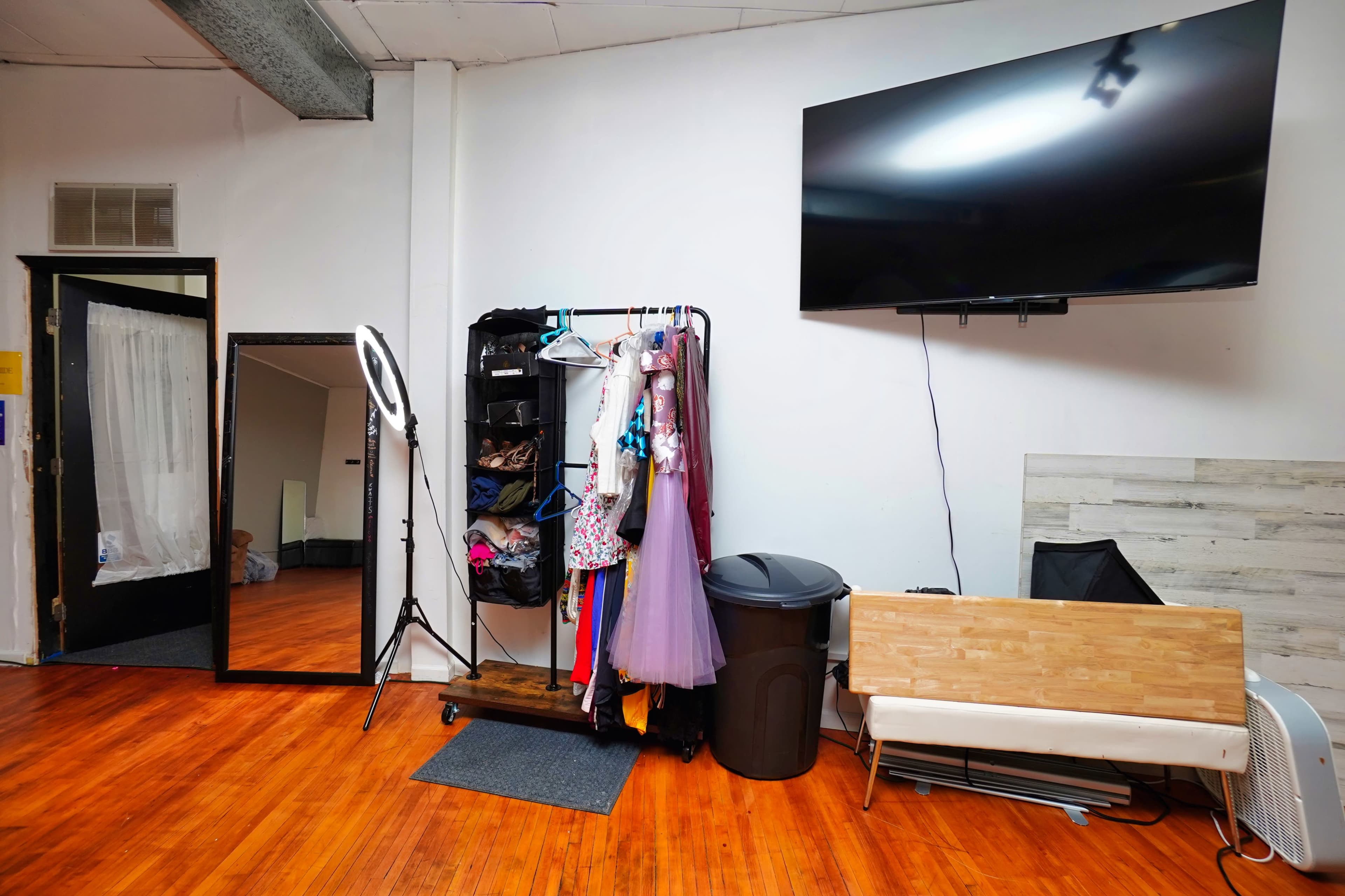 A room featuring a large flat-screen TV mounted on the wall, a clothing rack with various costumes, a full-length mirror, and a sitting area with a wooden bench.