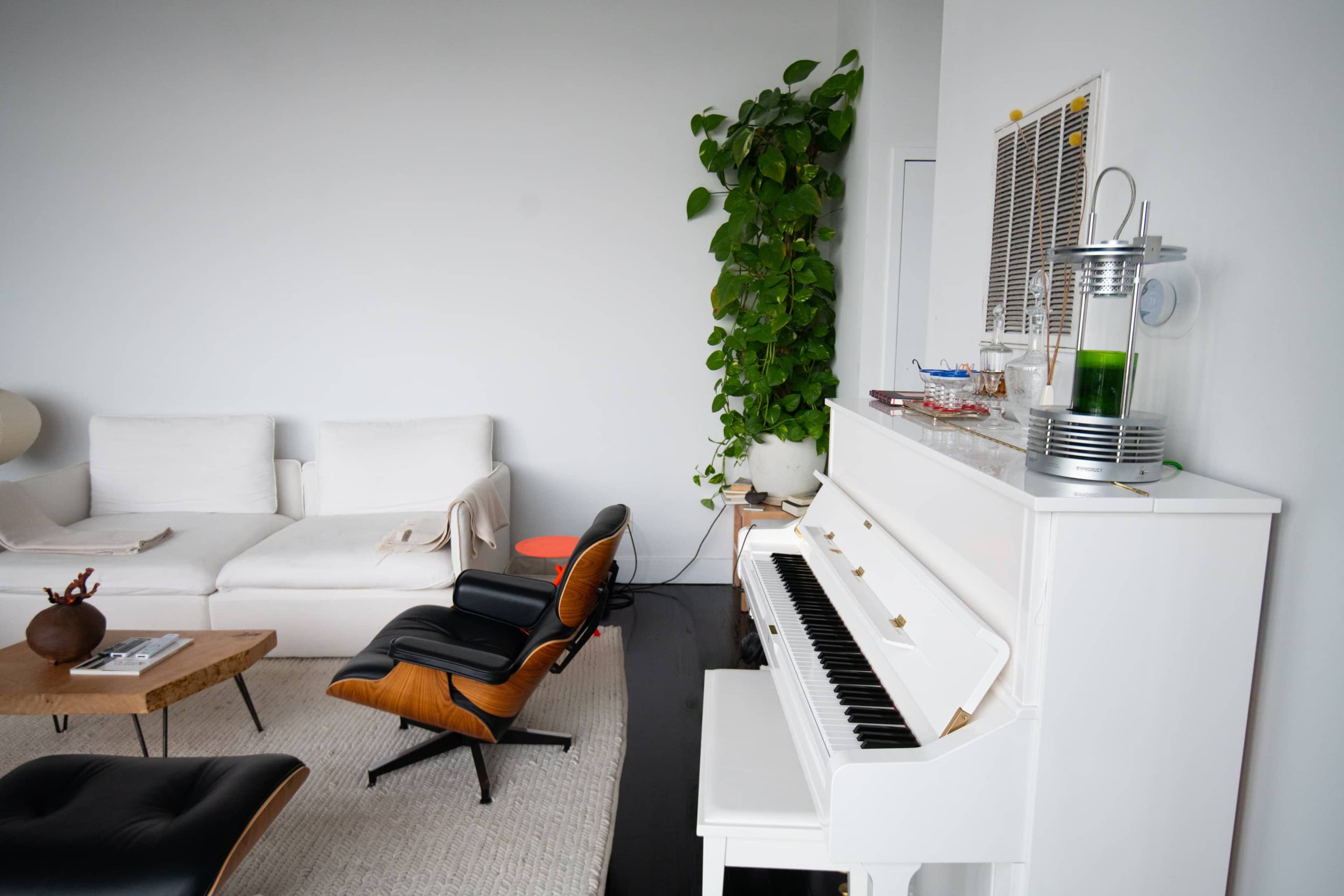 The scene features a white piano beside a modern black chair, a large green plant, and a light-colored sofa in a minimally furnished living room.