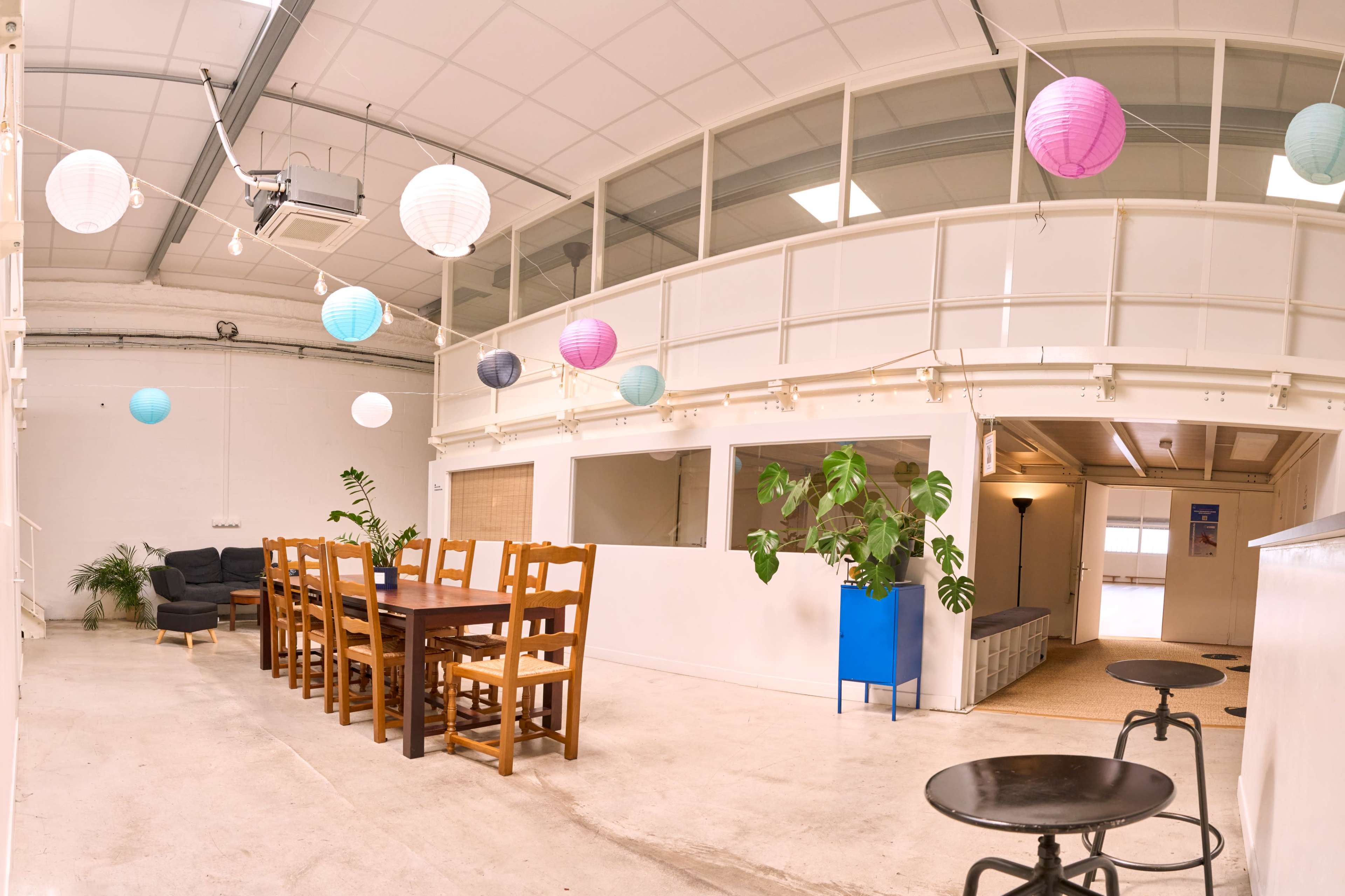 The image shows a spacious indoor area with a long wooden dining table surrounded by chairs, a green plant, and decorative paper lanterns hanging from the ceiling.