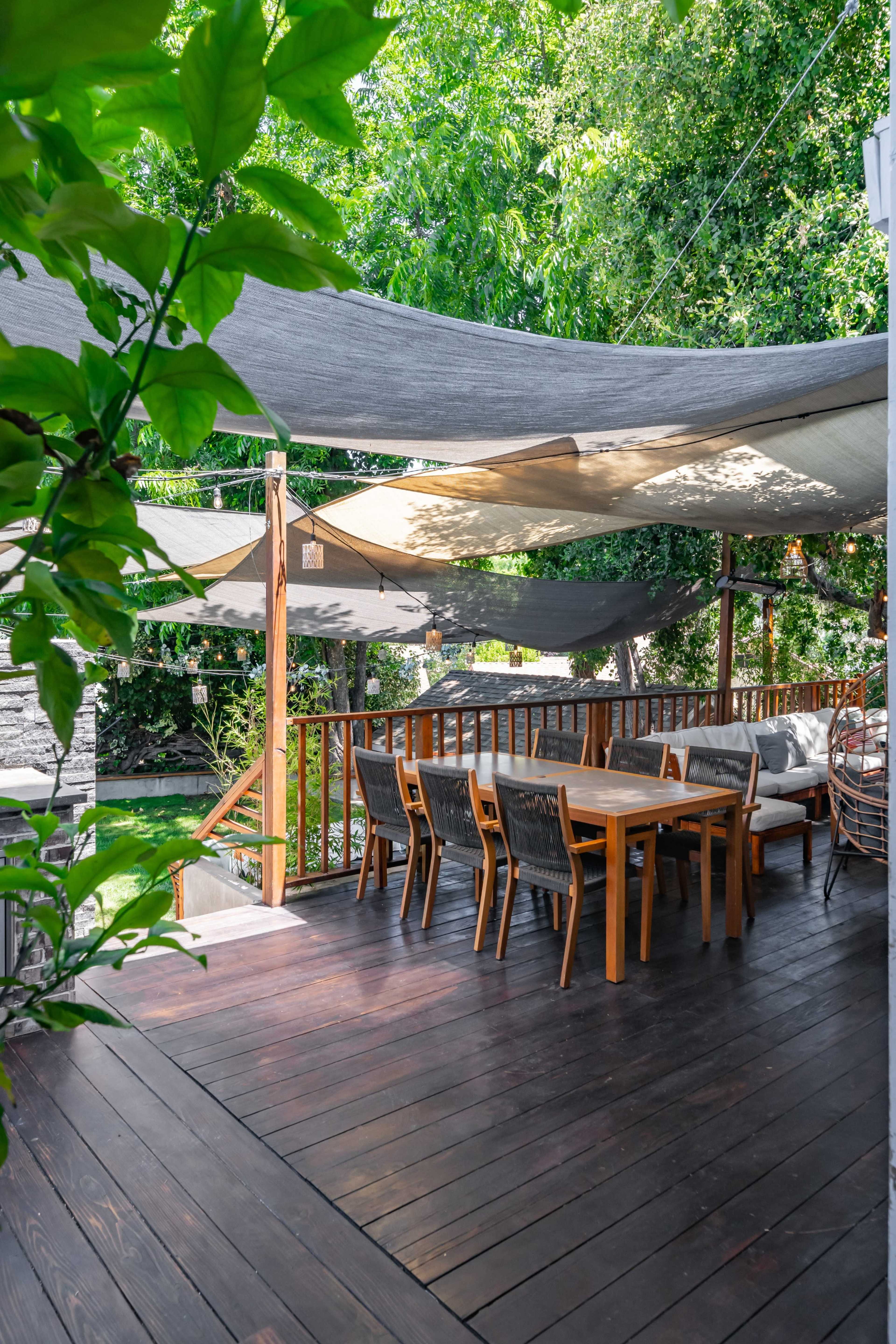 The image shows a wooden deck with a dining table and chairs, shaded by fabric canopies and surrounded by greenery.