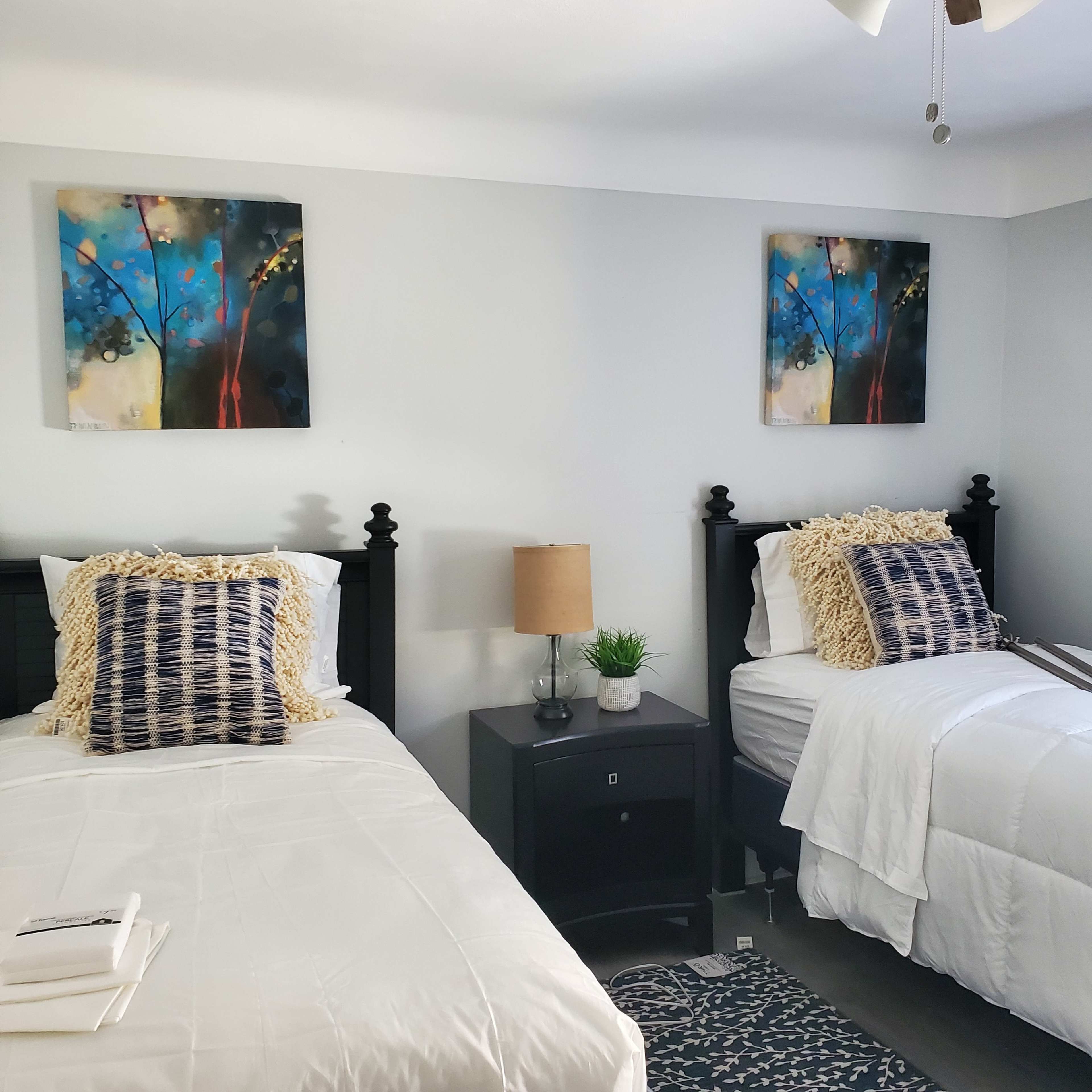 A brightly lit bedroom with two twin beds, each topped with patterned pillows, accompanied by a nightstand and abstract wall art.