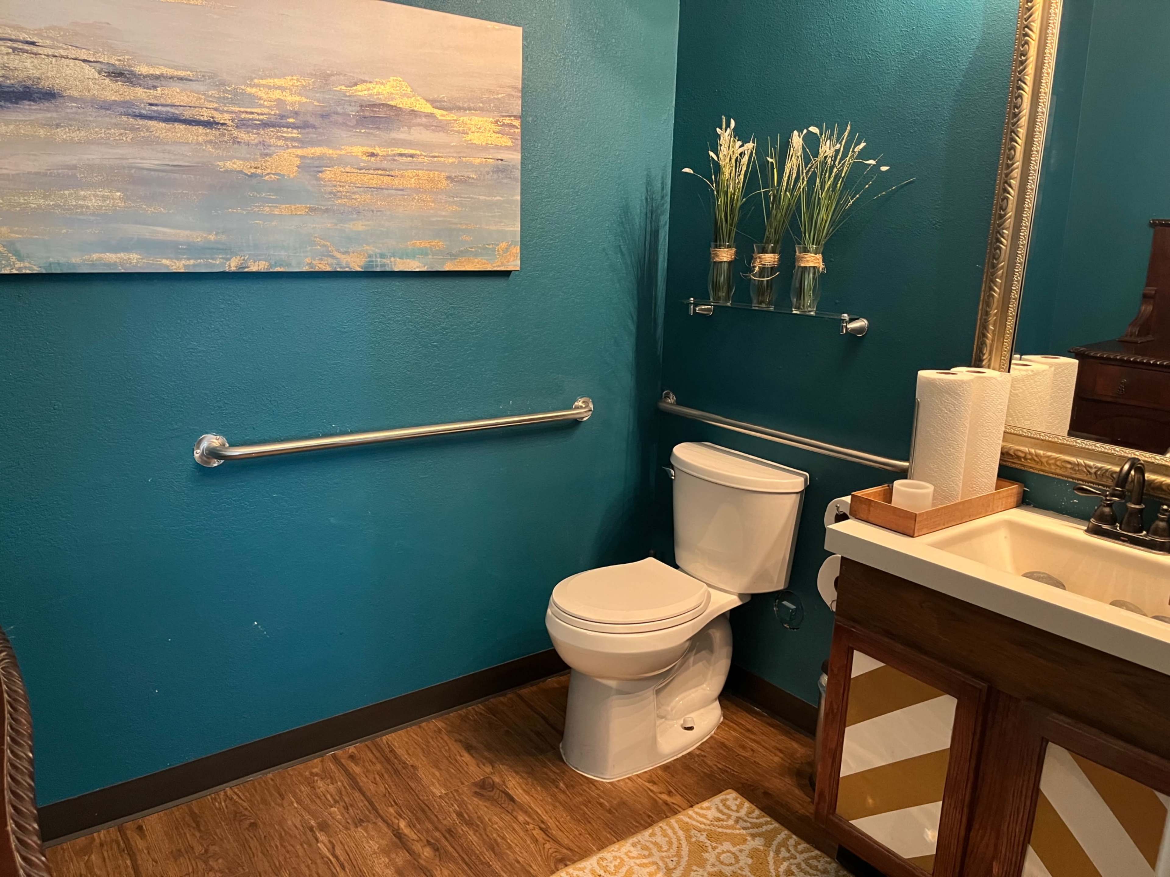 A bathroom with a teal-colored wall, a white toilet, a wooden vanity with a sink, and a framed painting above the sink.