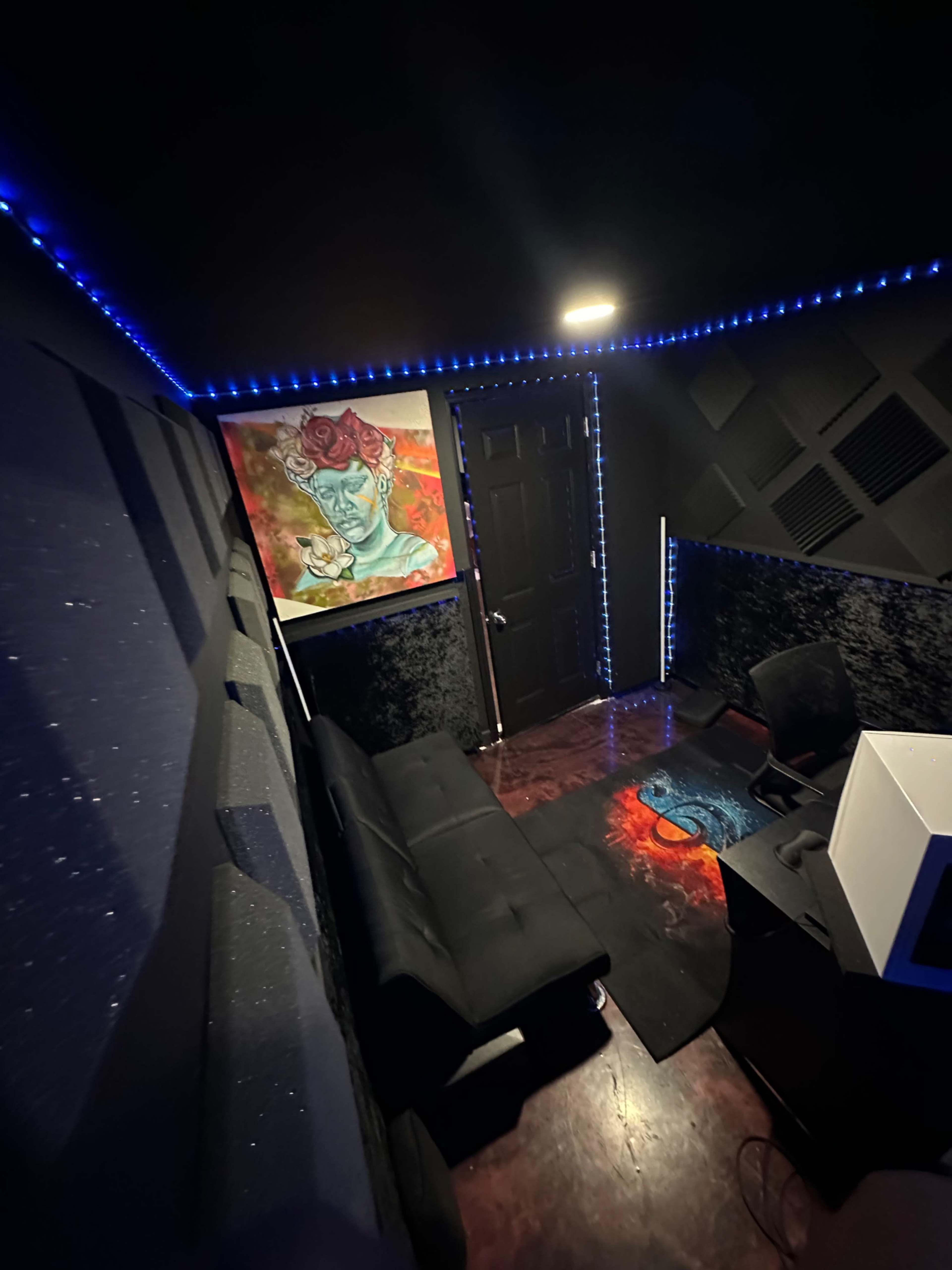 A dimly lit recording studio features a black couch, a desk with a computer, and a colorful piece of art on the wall, surrounded by soundproof panels and LED lighting.