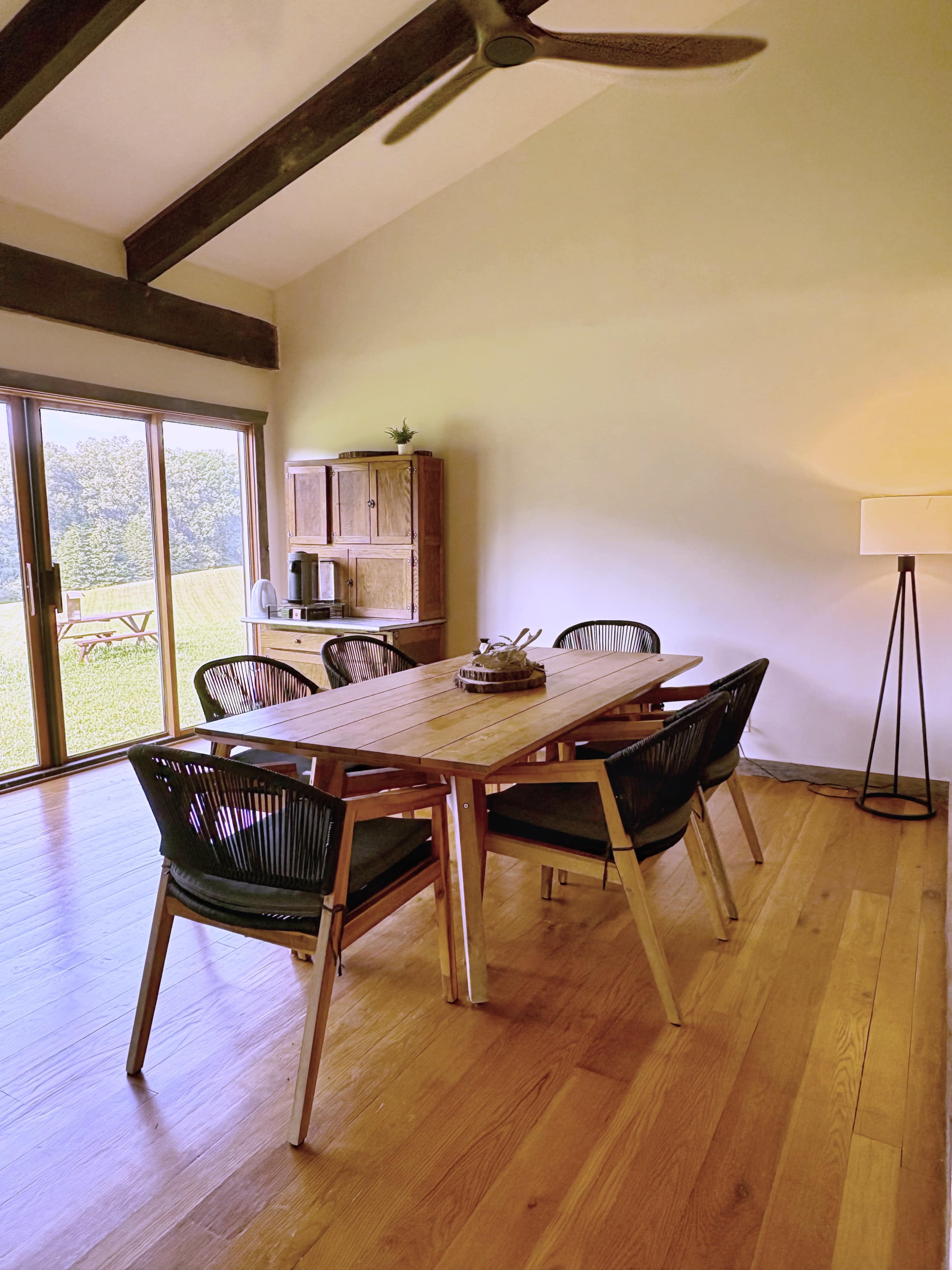 The image shows a modern dining area with a wooden table surrounded by six chairs, large windows letting in natural light, and a view of greenery outside.