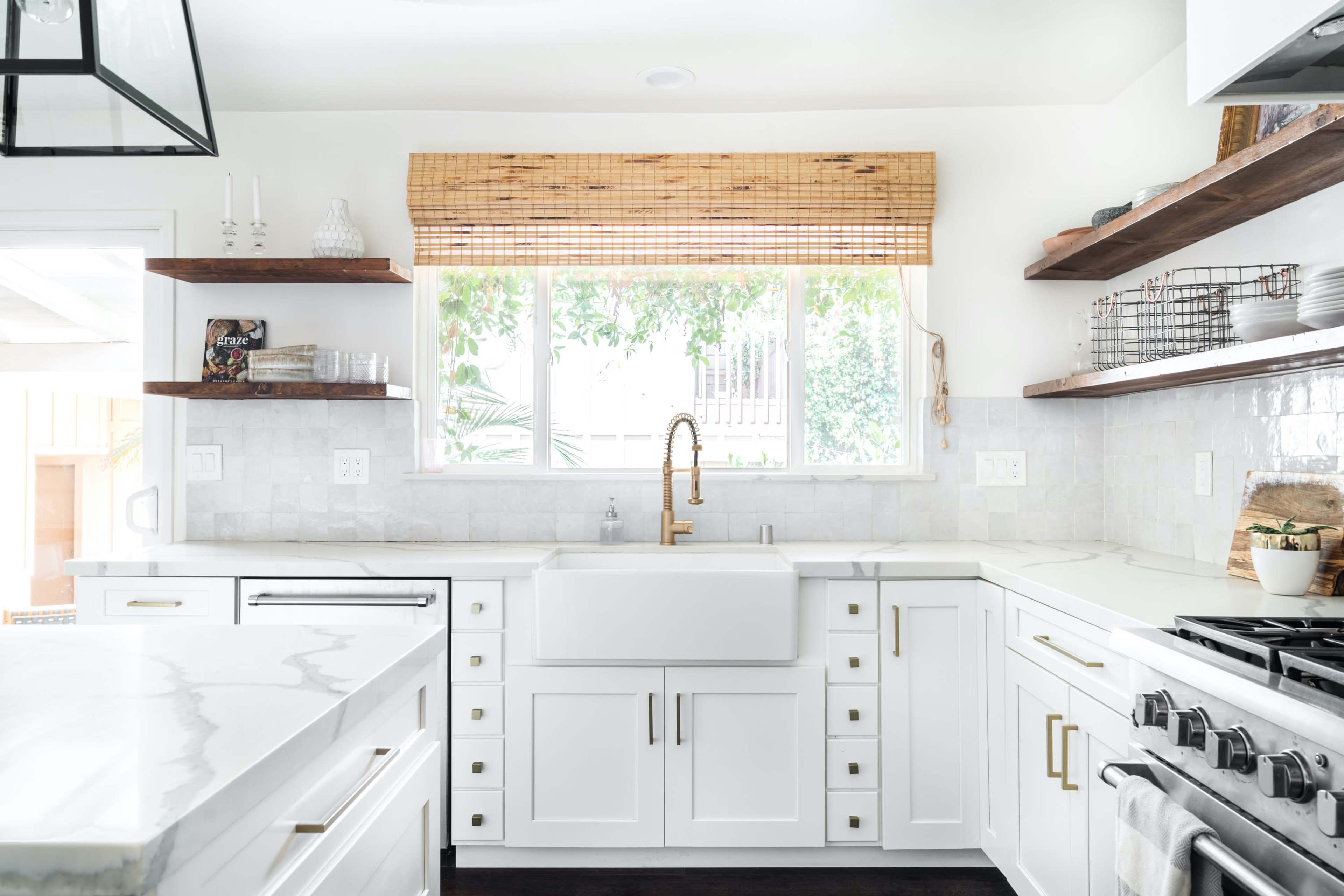 A modern kitchen with white cabinets, a large farmhouse sink, and wooden shelves, illuminated by natural light from a window.