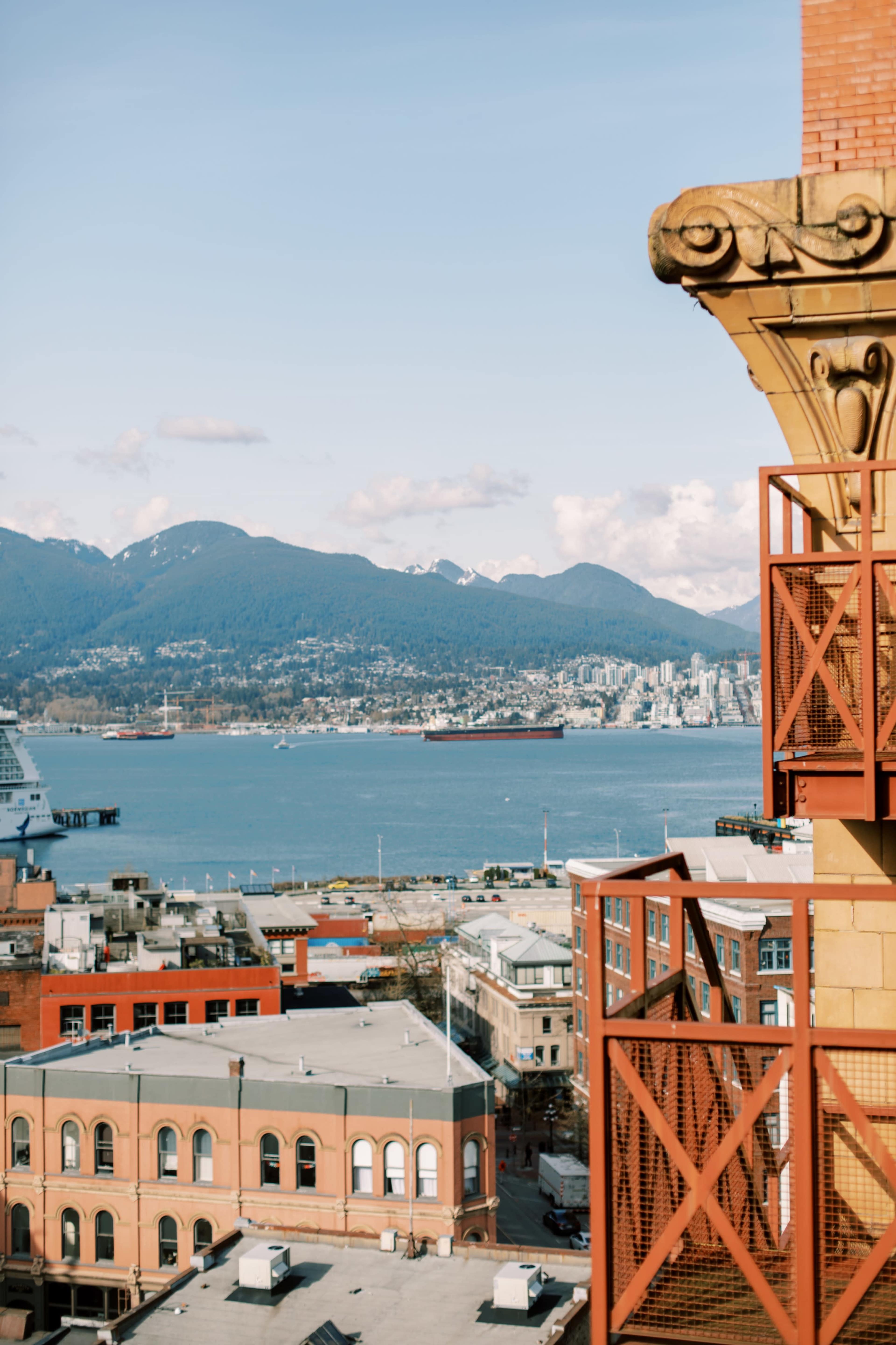 A view of a coastal city with mountains in the background, featuring a prominent building corner and a harbor with ships.