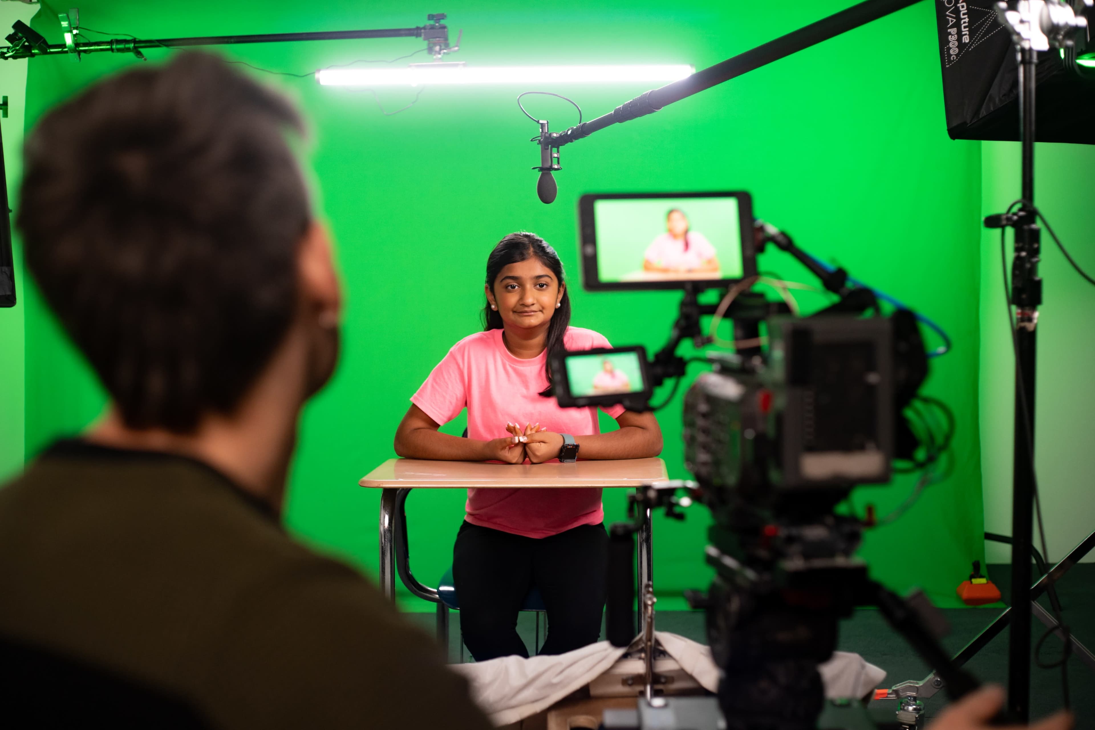 A young woman sits at a desk in front of a green screen, while a camera operator adjusts settings on a camera recording her.